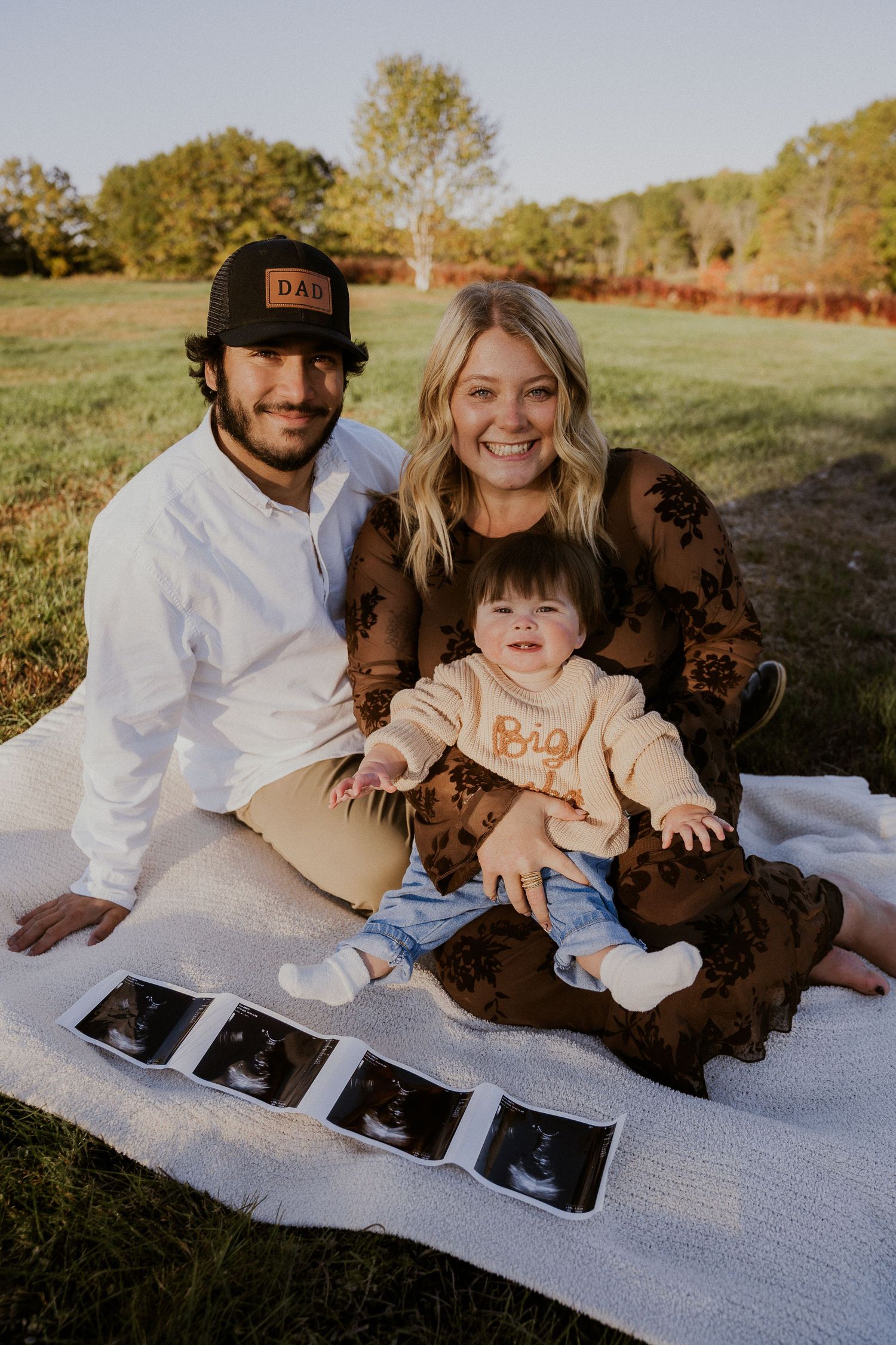 Family sitting on blanket outdoors with ultrasound photos and a small child.