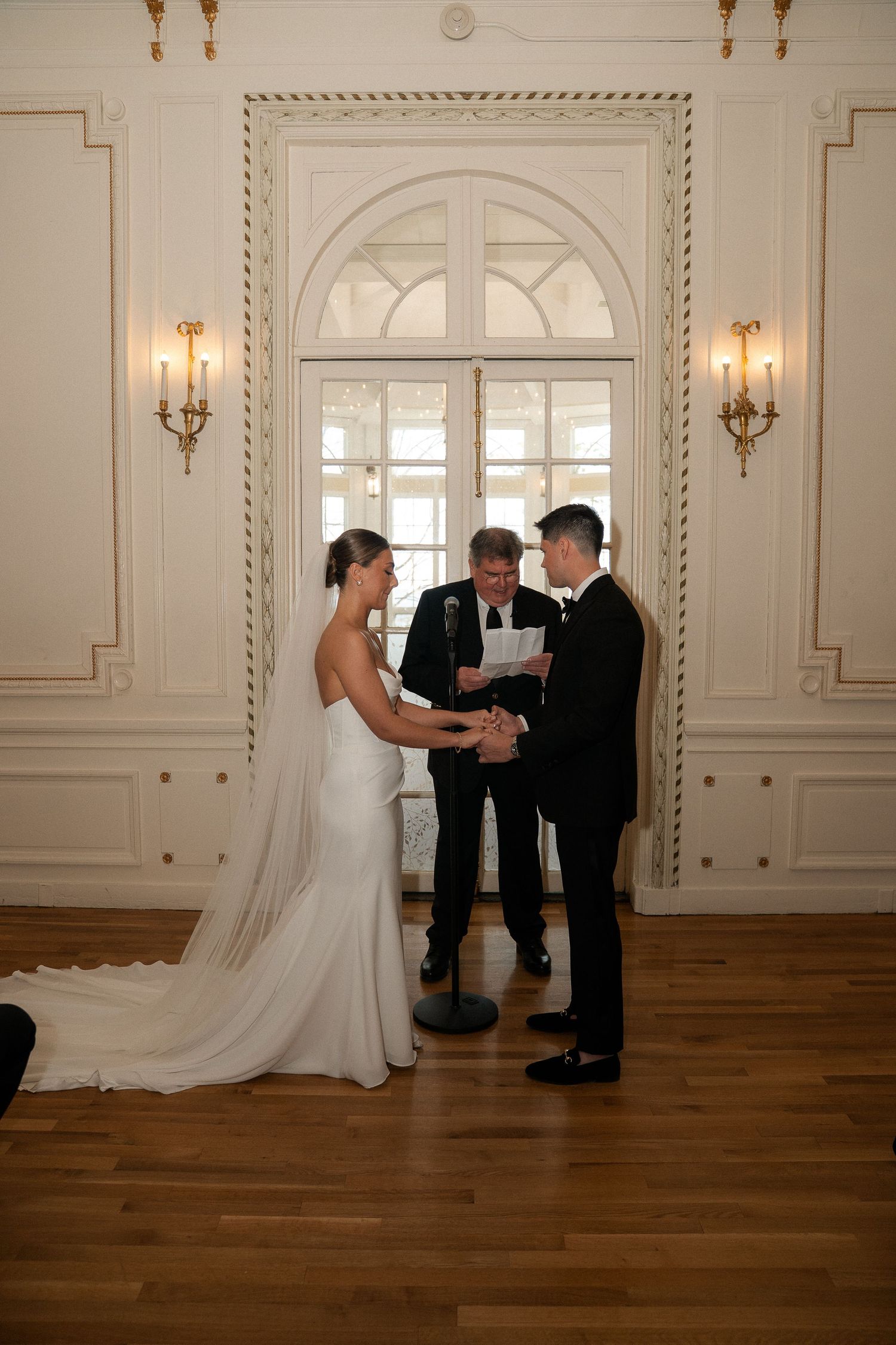 Bride and groom exchange vows before an officiant in an elegant white ballroom during their wedding ceremony.