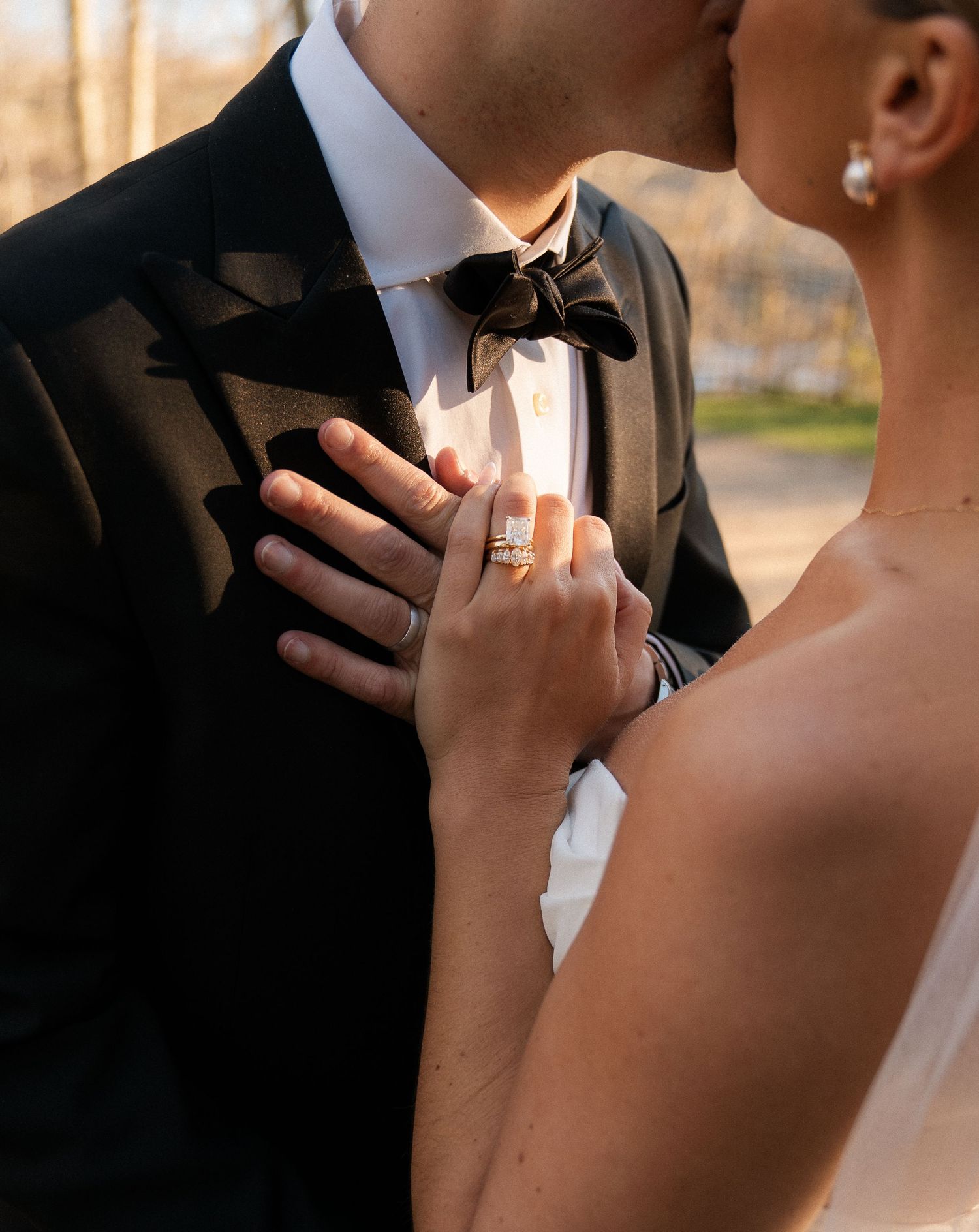 Close-up of bride's hand with diamond engagement ring resting on groom's black tuxedo lapel.