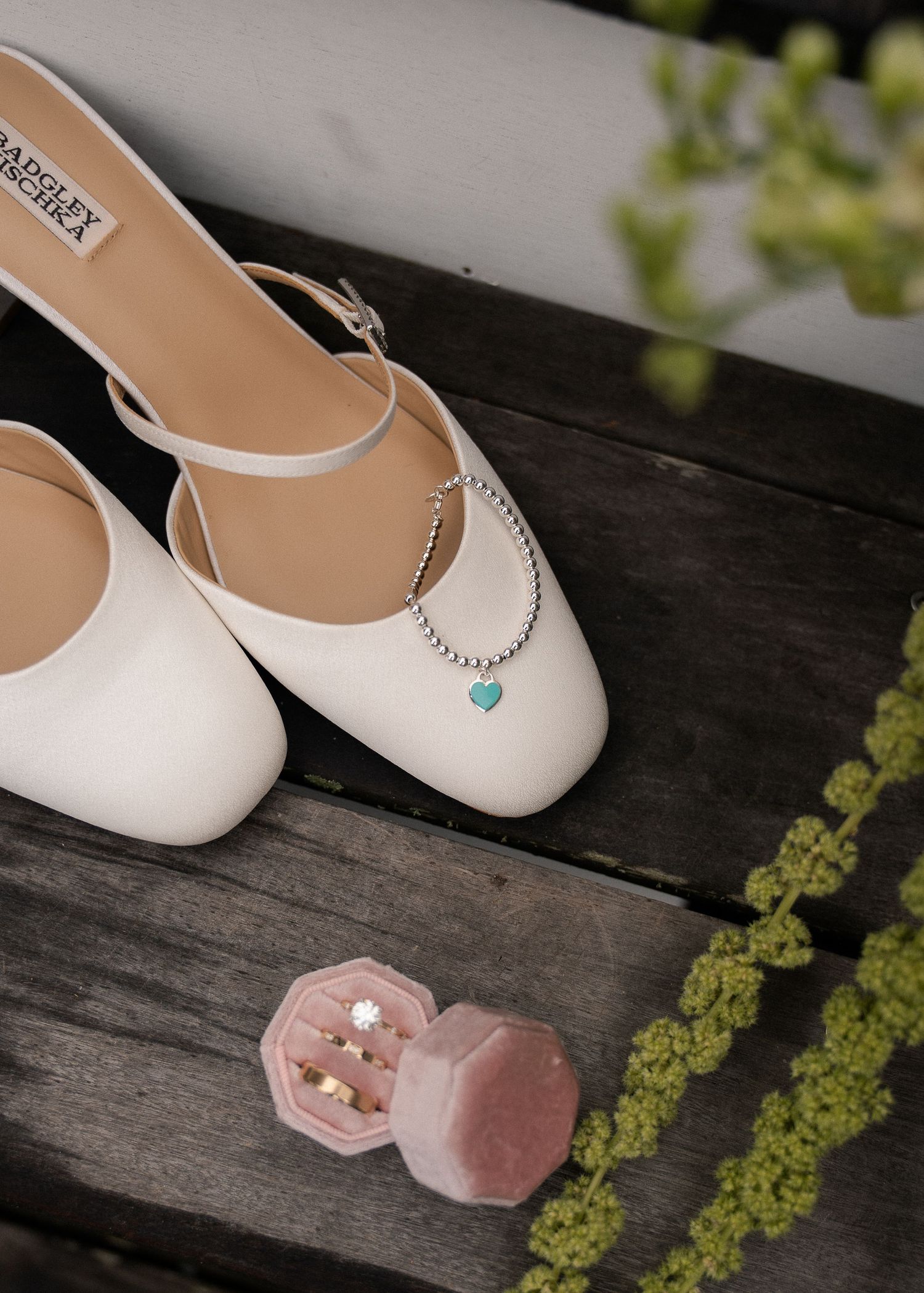 White bridal heels with pearl bracelet and turquoise heart charm beside pink velvet ring box with gold rings.