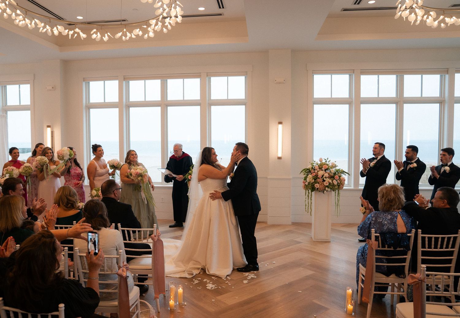 Bride and groom share first kiss at elegant waterfront wedding ceremony with floor-to-ceiling windows and floral arrangements.
