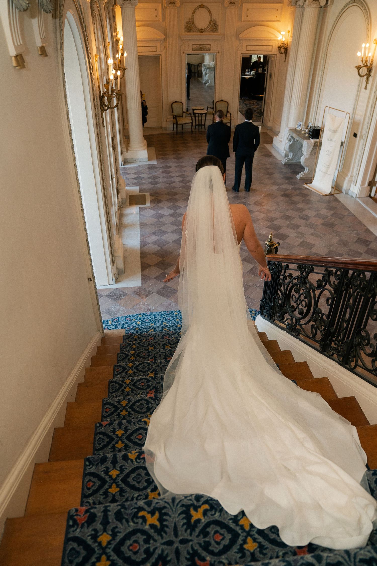 Bride in flowing white gown and veil descends ornate staircase in elegant venue toward groom.