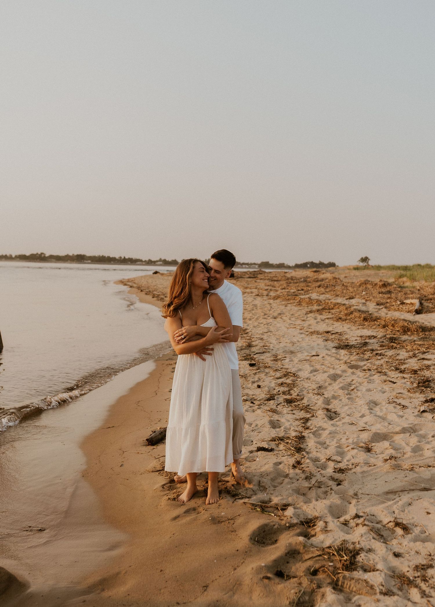 A romantic couple embraces on a sandy beach at sunset wearing white and beige outfits.