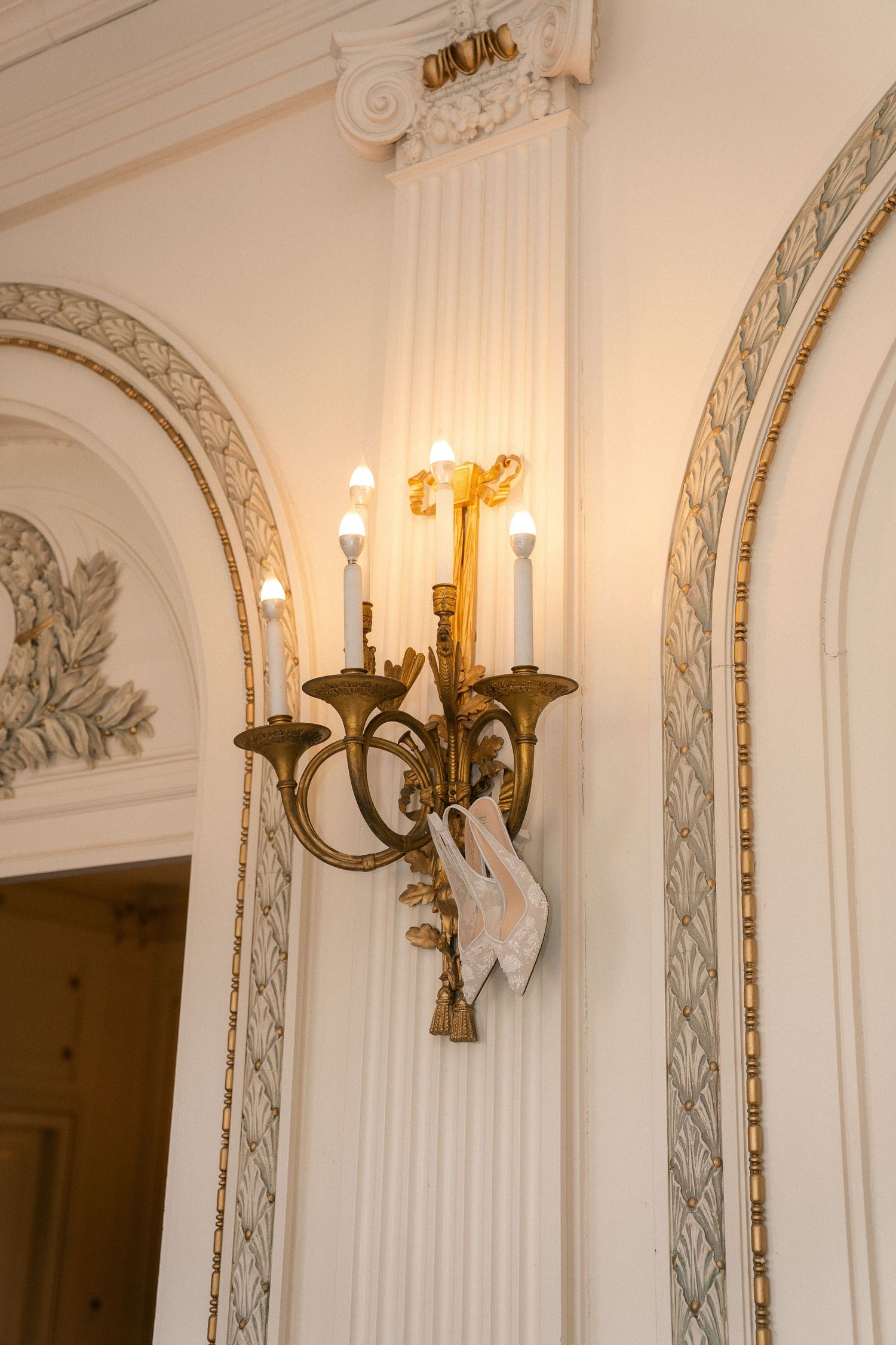 Ornate gold wall sconce with lit candles and white bridal shoes hung on it, beside elegant arched molding.
