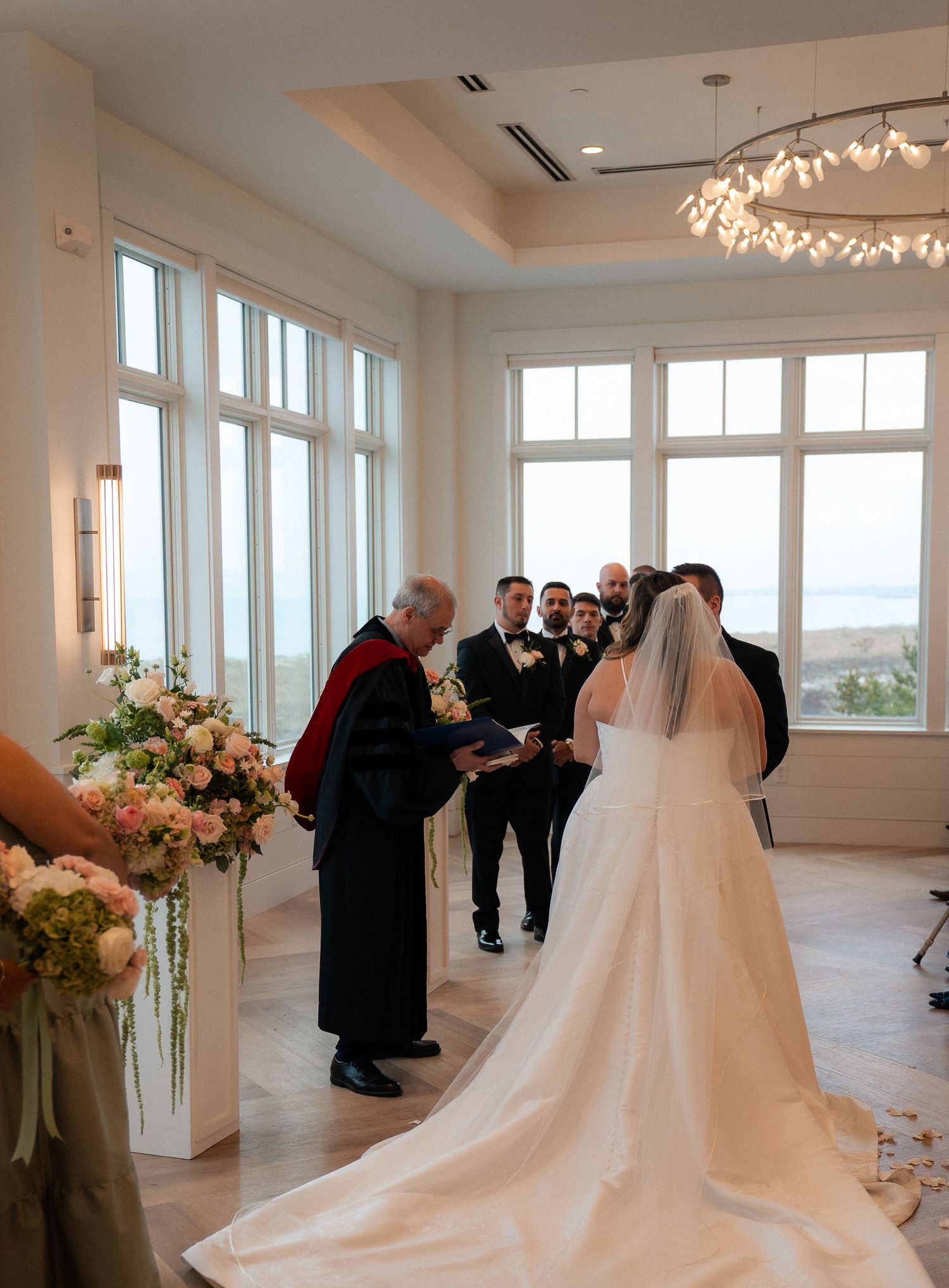 Bride and groom exchange vows at elegant indoor wedding ceremony with large windows and floral arrangements.