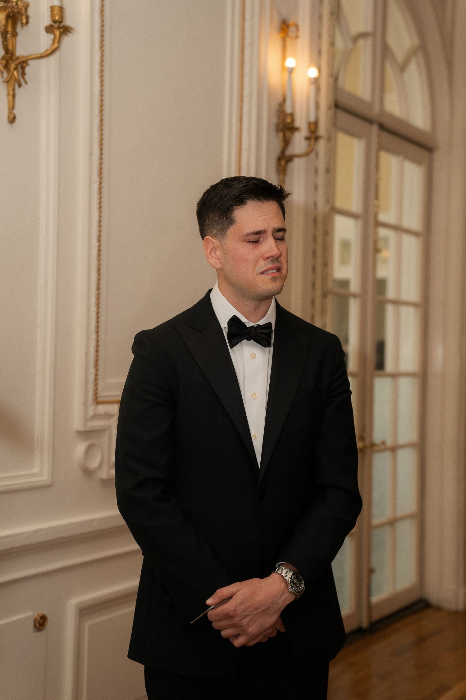 Emotional groom in black tuxedo and bow tie stands in elegant white room with gold wall sconces.