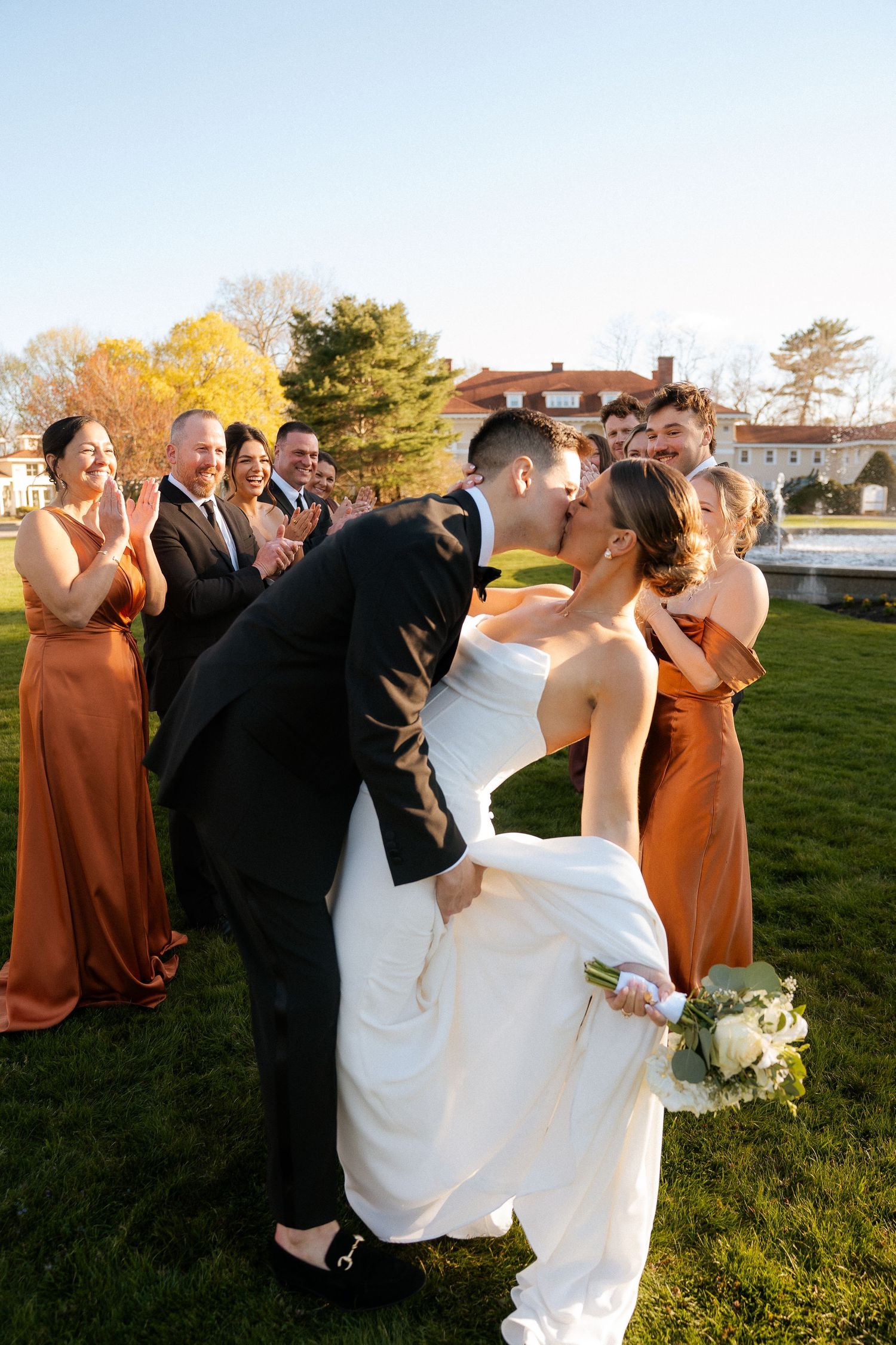 Bride and groom share a romantic dip kiss surrounded by cheering wedding party in rust bridesmaid dresses on a sunny lawn.