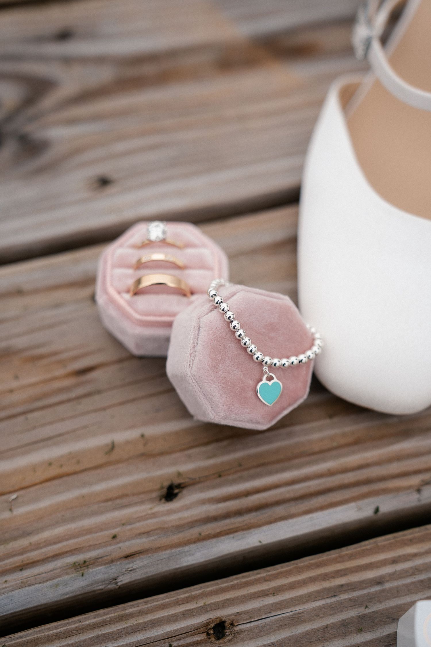 Pink velvet ring box with wedding bands and diamond ring beside a silver heart charm bracelet and white bridal heel.