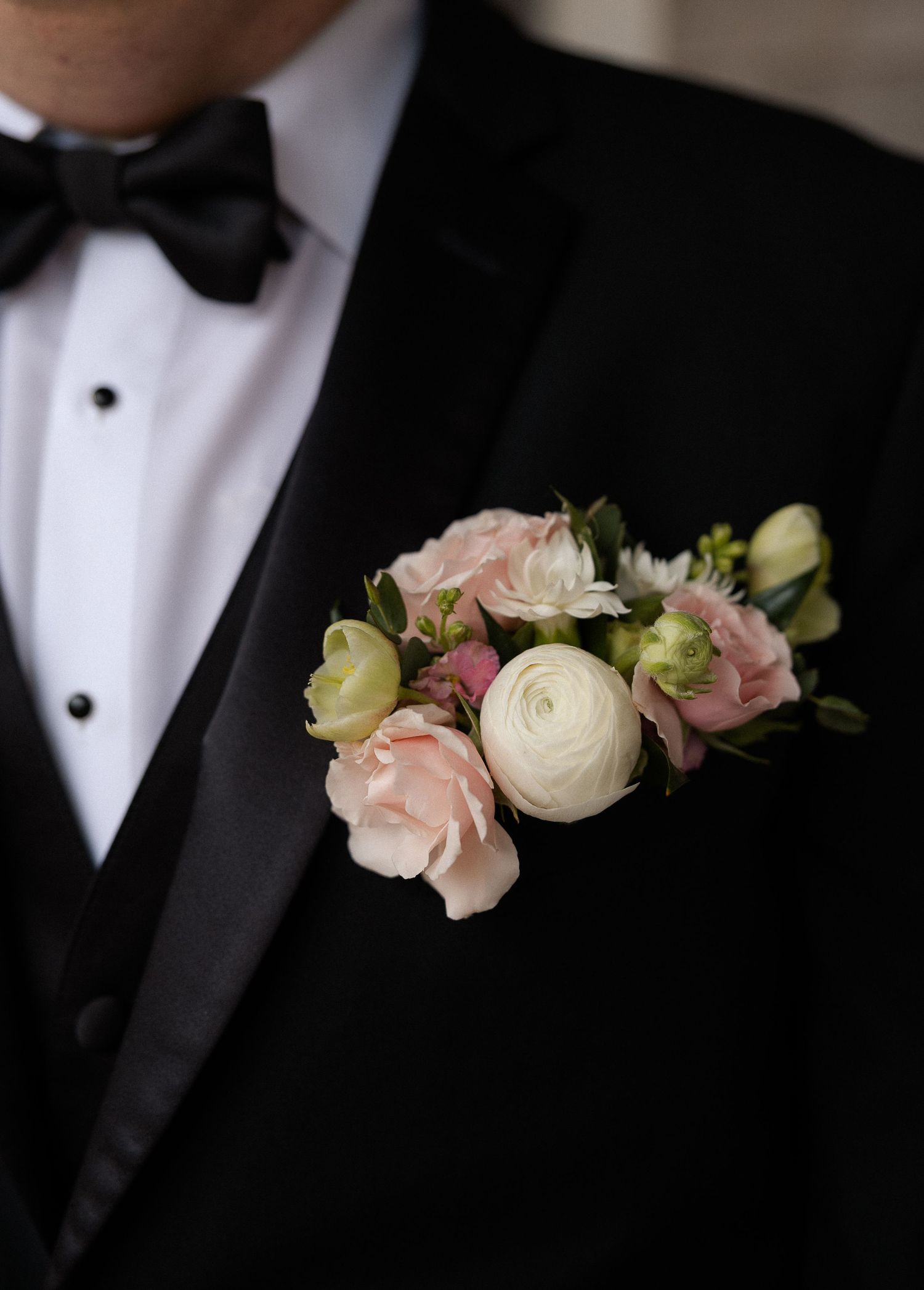 Close-up of elegant pink and white ranunculus boutonniere on groom's black tuxedo jacket with bow tie.
