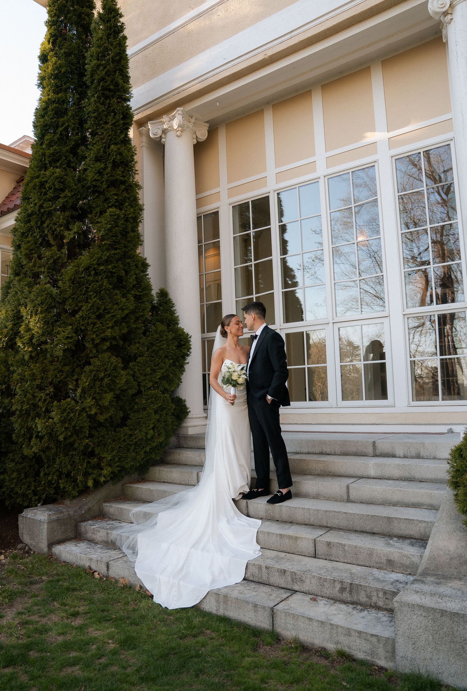 Bride and groom share a kiss on stone steps beside tall cypress trees at wedding venue.