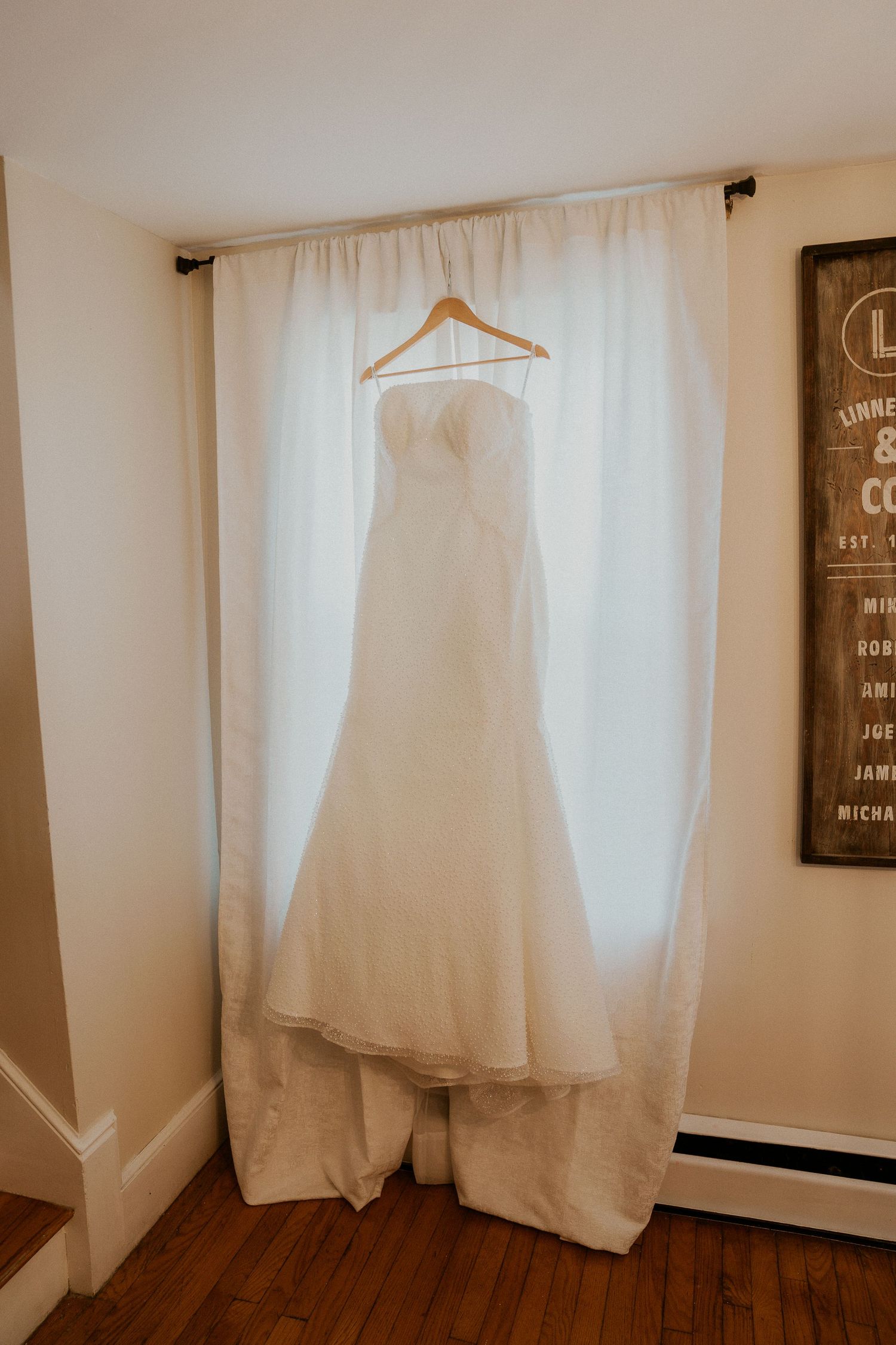 A white wedding dress hangs elegantly against a neutral wall in soft, natural lighting.