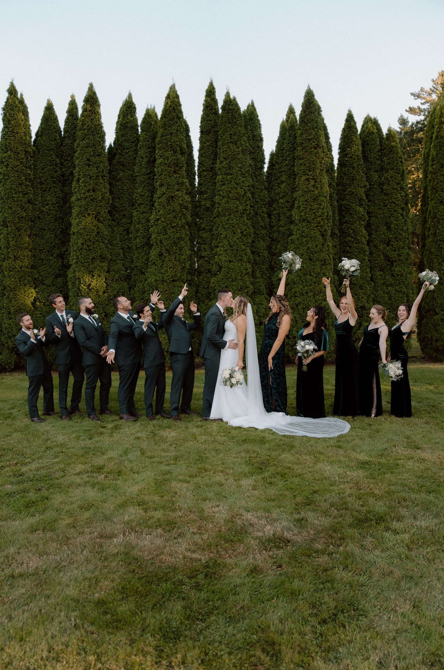 Wedding party celebrates with raised bouquets against a wall of cypress trees.