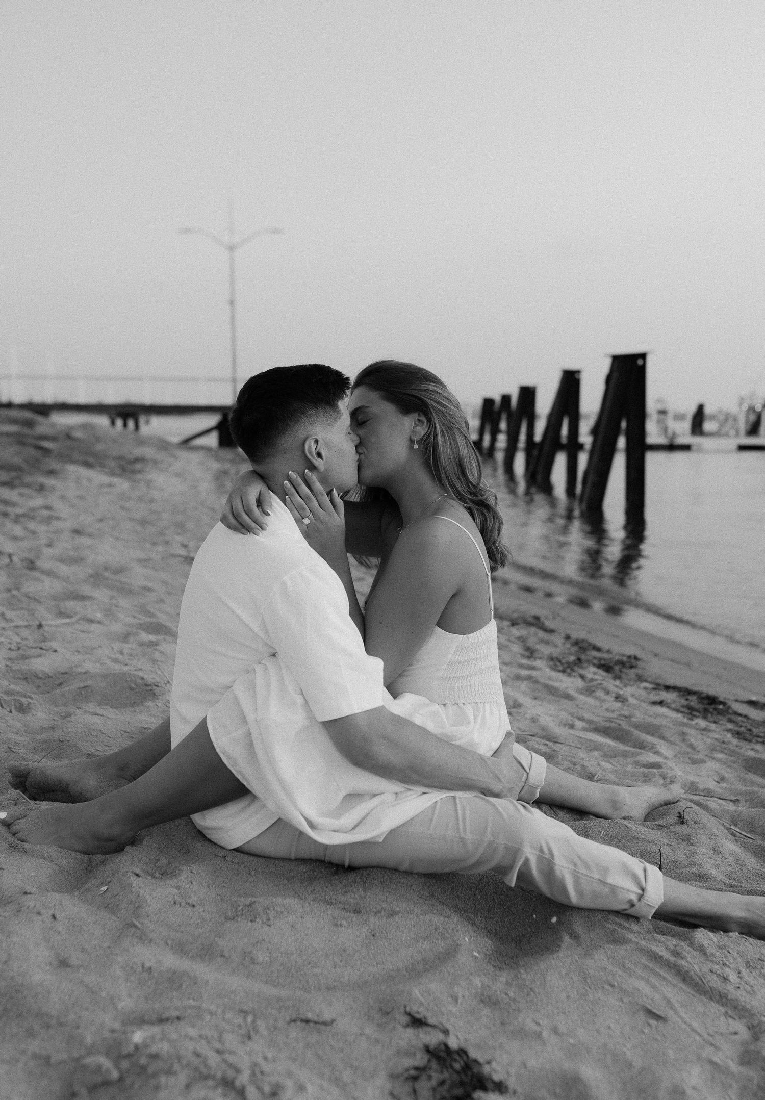 Black and white photo of couple sharing intimate moment on beach with pier silhouette.