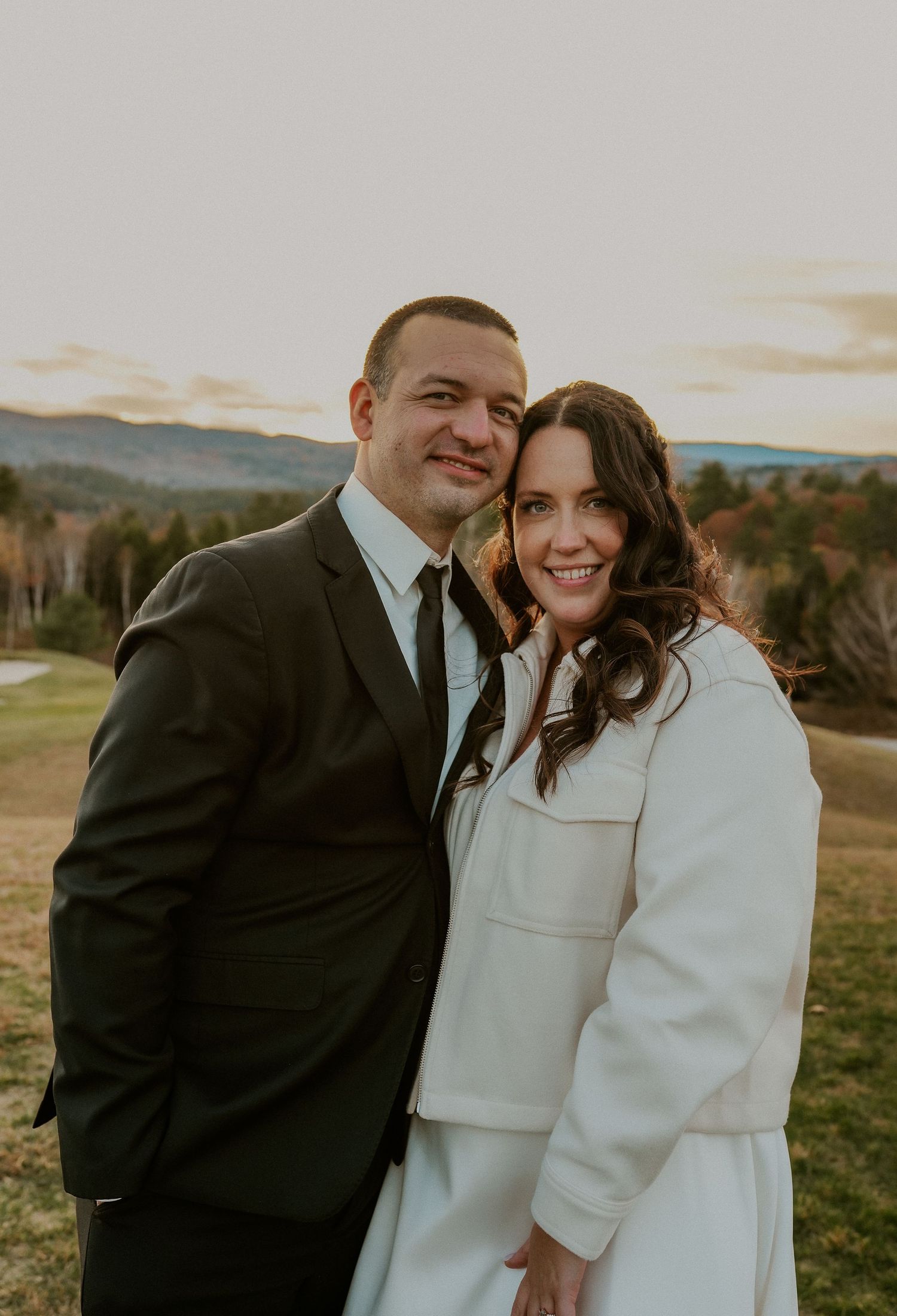 A couple poses outdoors in wedding attire with mountains and autumn foliage in the background.