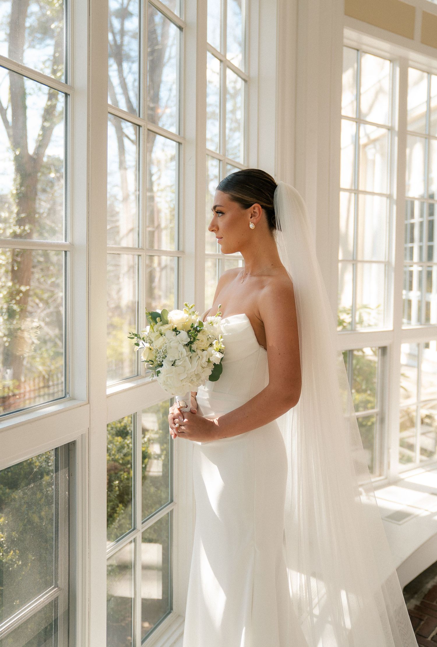Elegant bride in strapless white gown holding bouquet, standing by large windows with natural light.