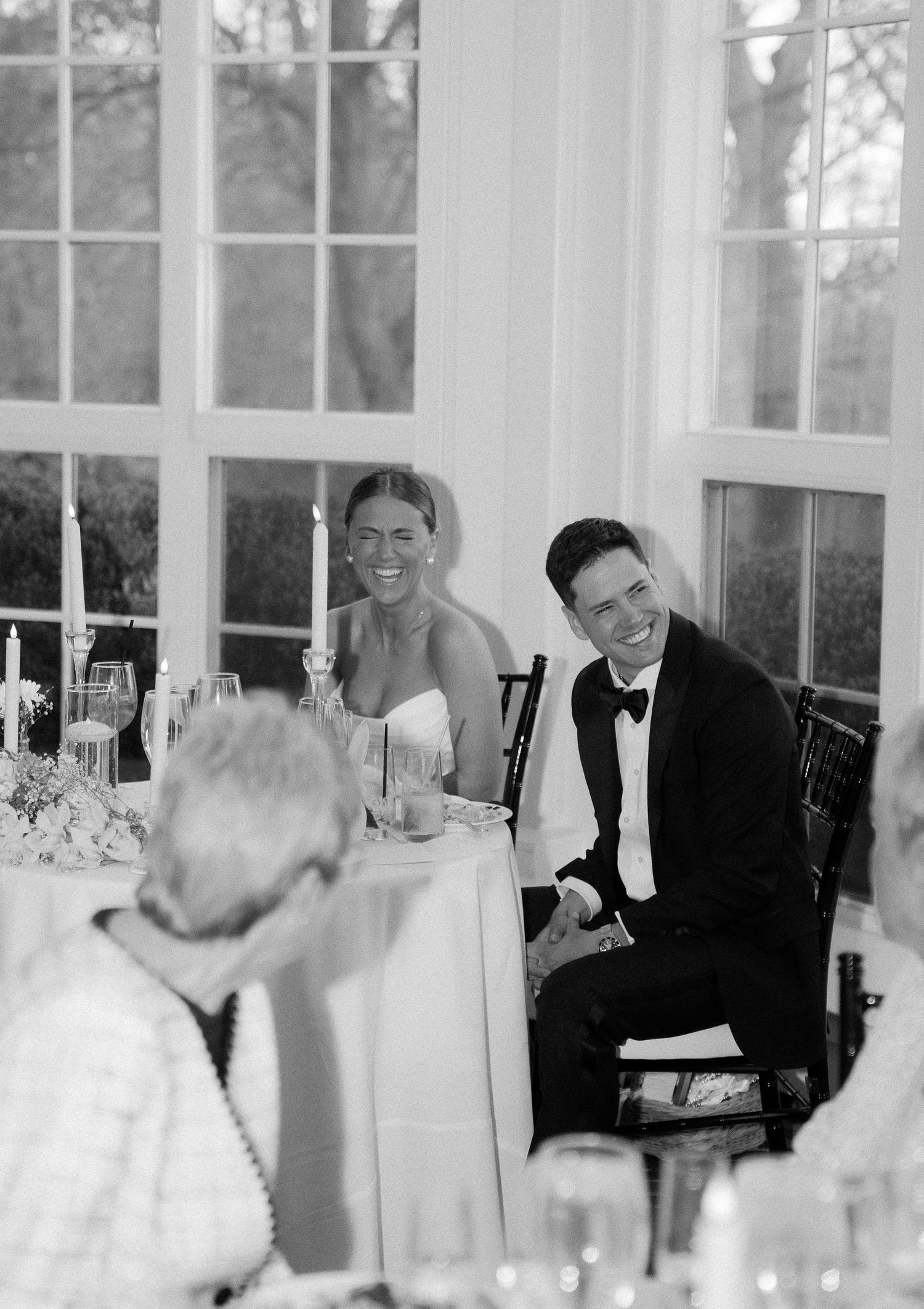 Black and white photo of smiling bride and groom seated at sweetheart table during wedding reception.
