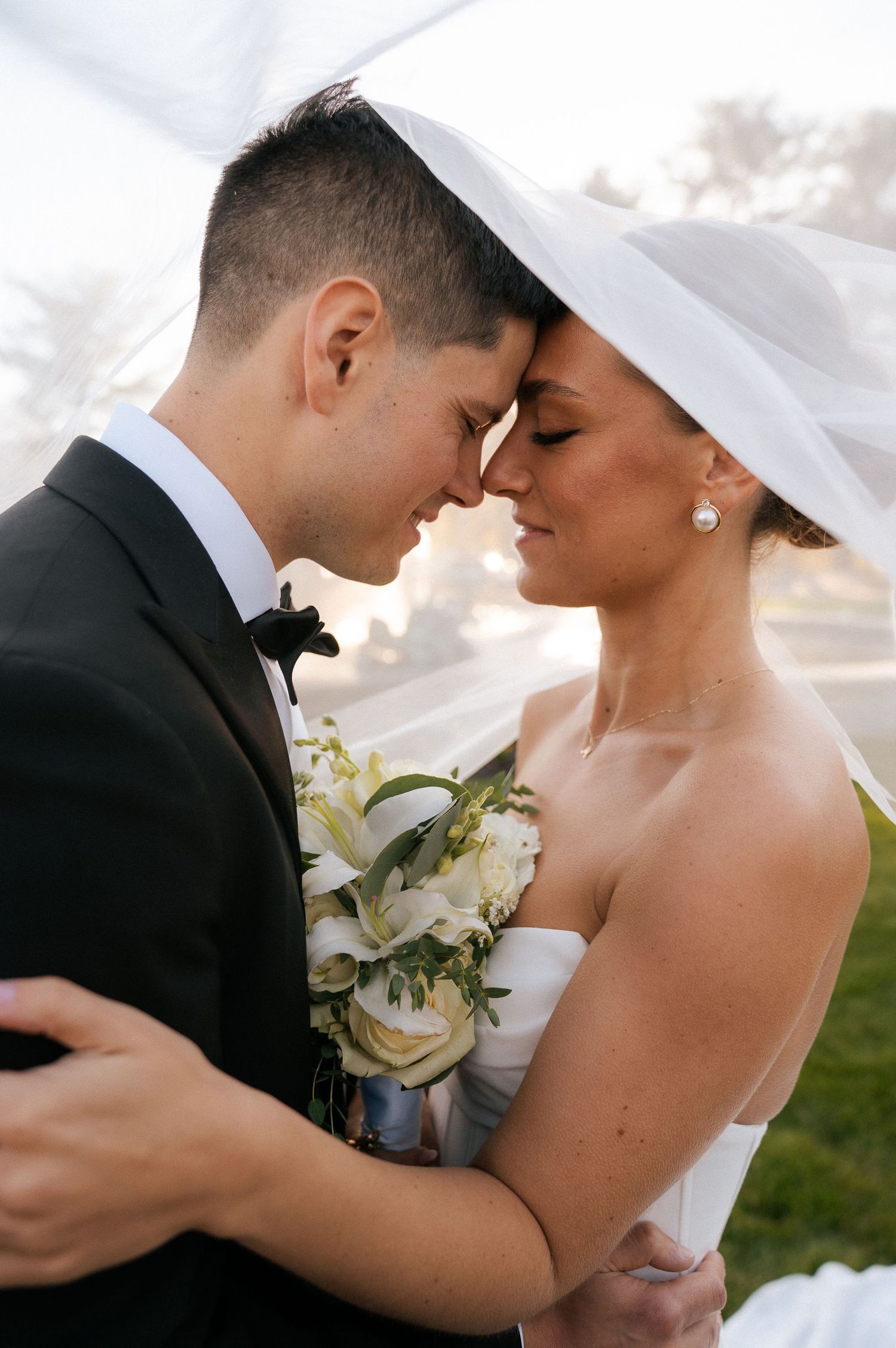 Bride and groom touching foreheads under wedding veil, sharing intimate moment with white floral bouquet.