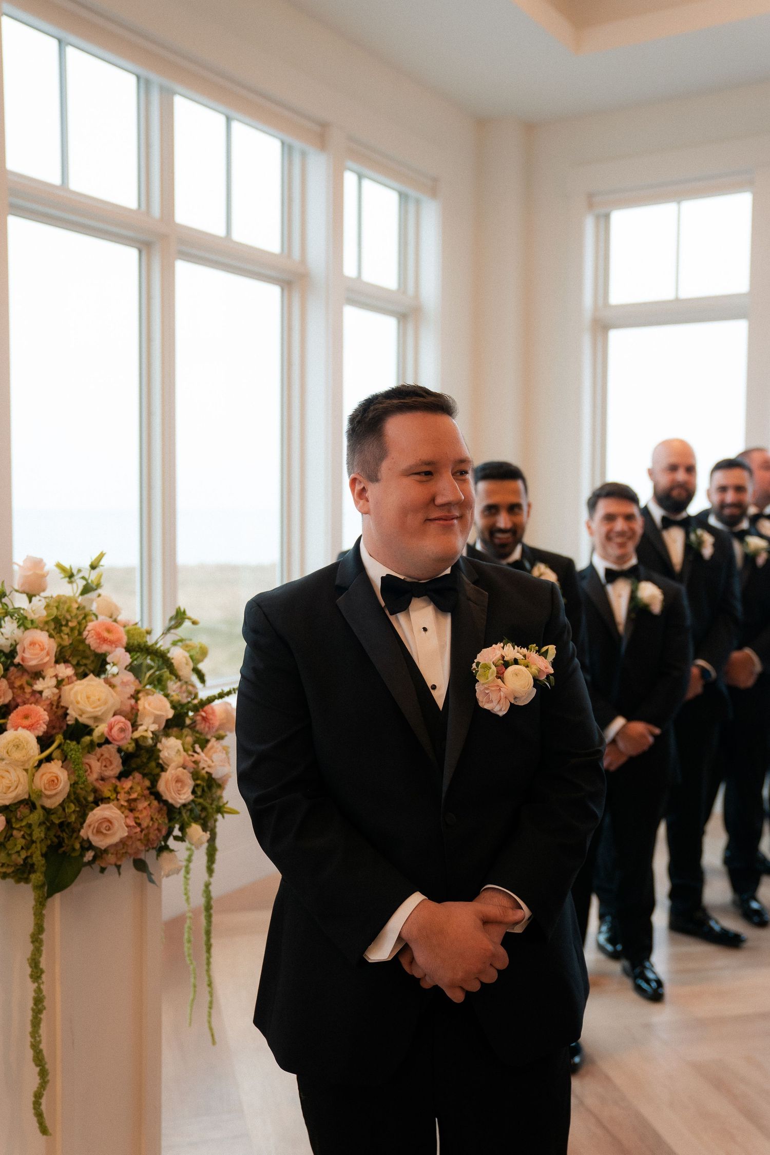 Groom in black tuxedo smiling at altar with groomsmen lined up behind him during wedding ceremony.