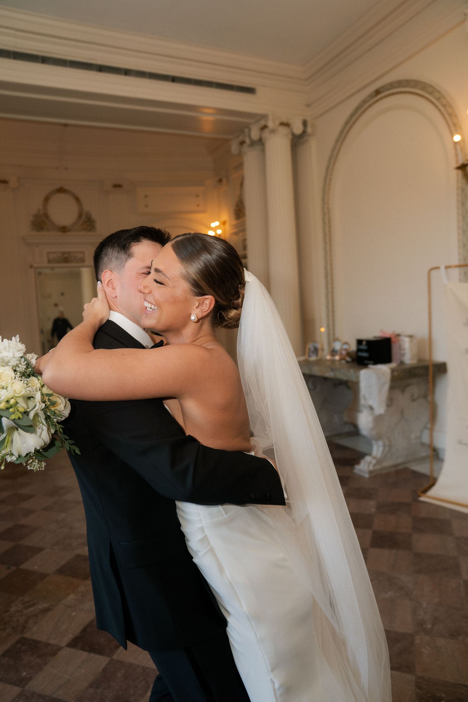 Radiant bride and groom share a warm embrace while smiling in an elegant historic wedding venue.