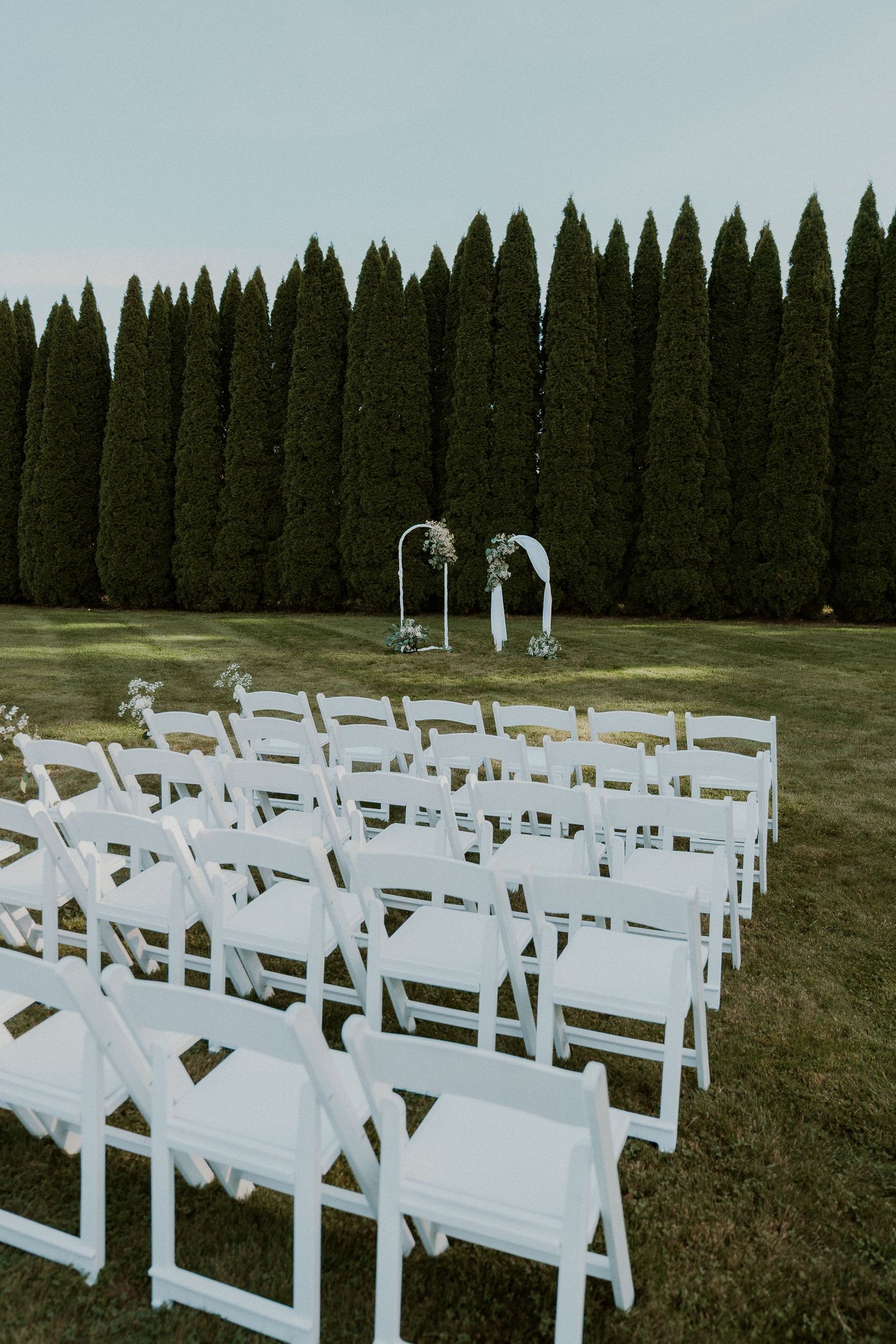 White ceremony chairs arranged outdoors with evergreen trees creating a natural backdrop.