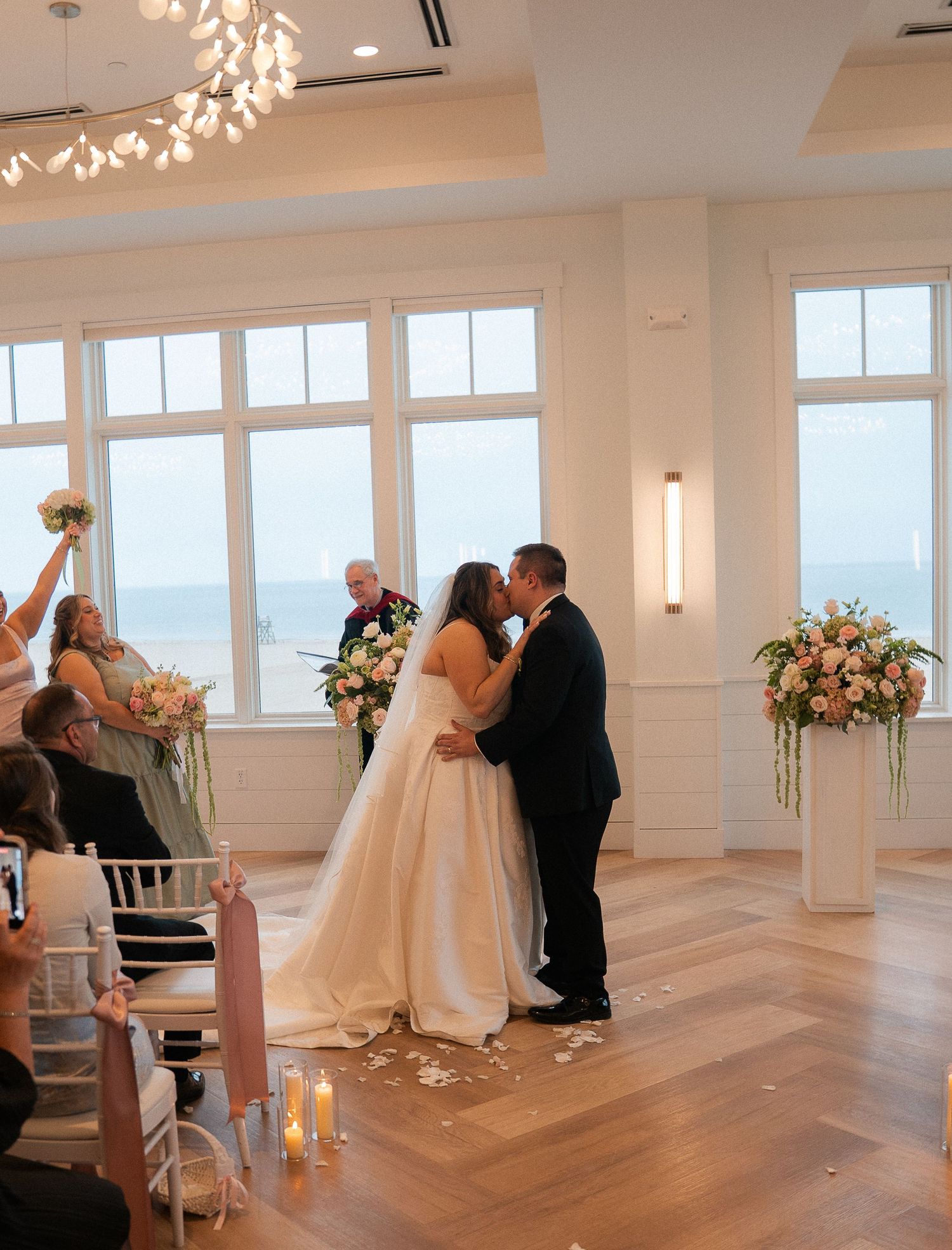 Bride and groom share first kiss at altar during elegant indoor wedding ceremony with ocean backdrop.
