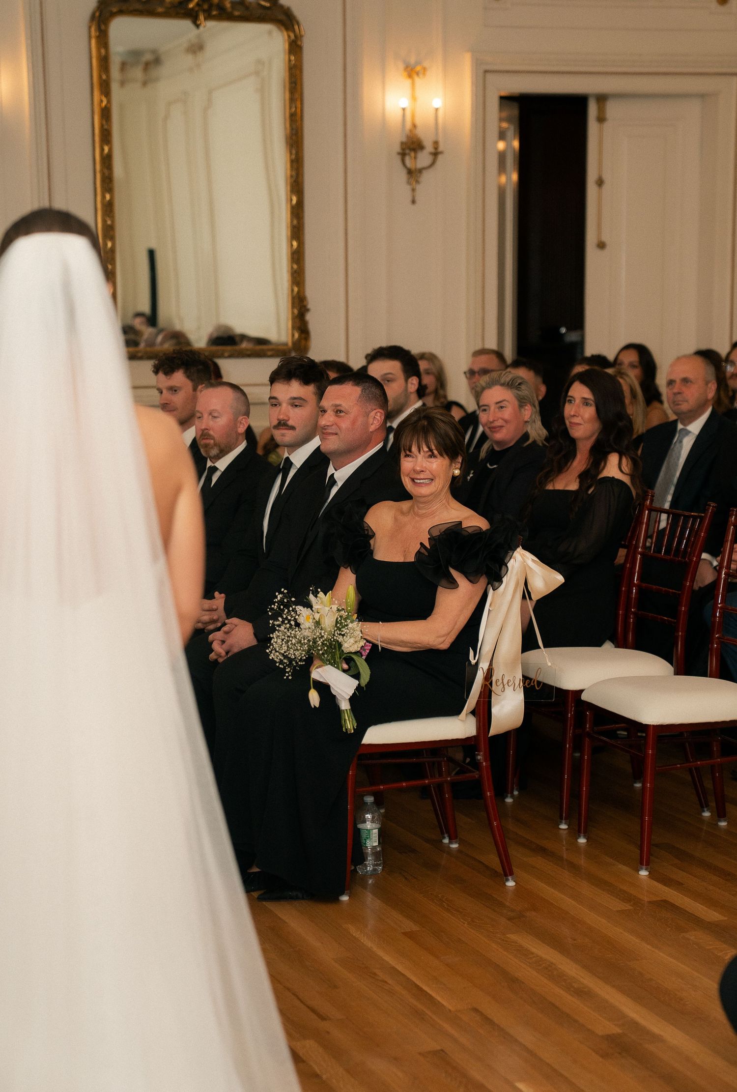 Wedding guests in black attire smile warmly during a ceremony, seated in rows with a bride in white veil visible in foreground.