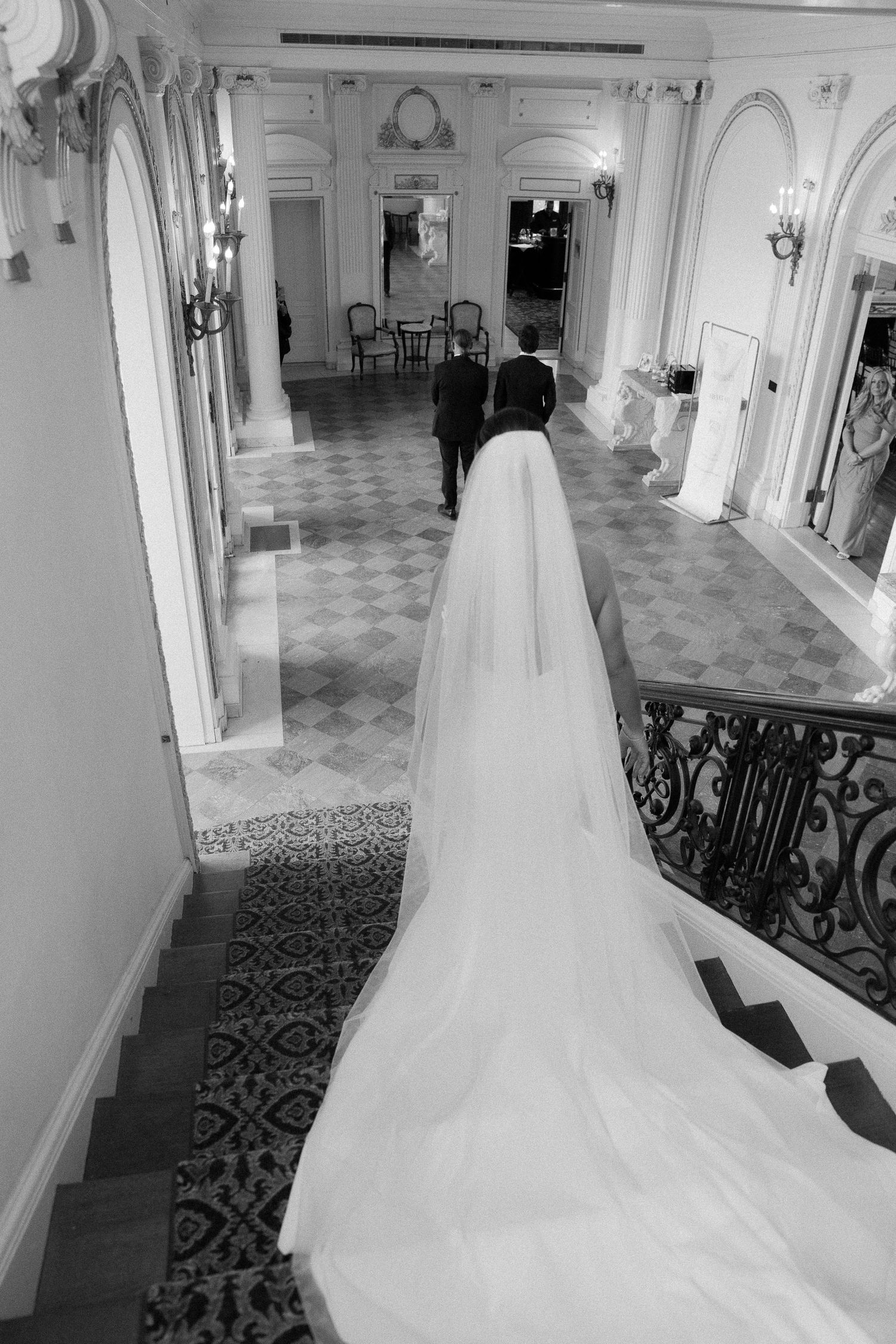 Black and white photo of bride in flowing cathedral veil descending ornate staircase with groom in classic venue.