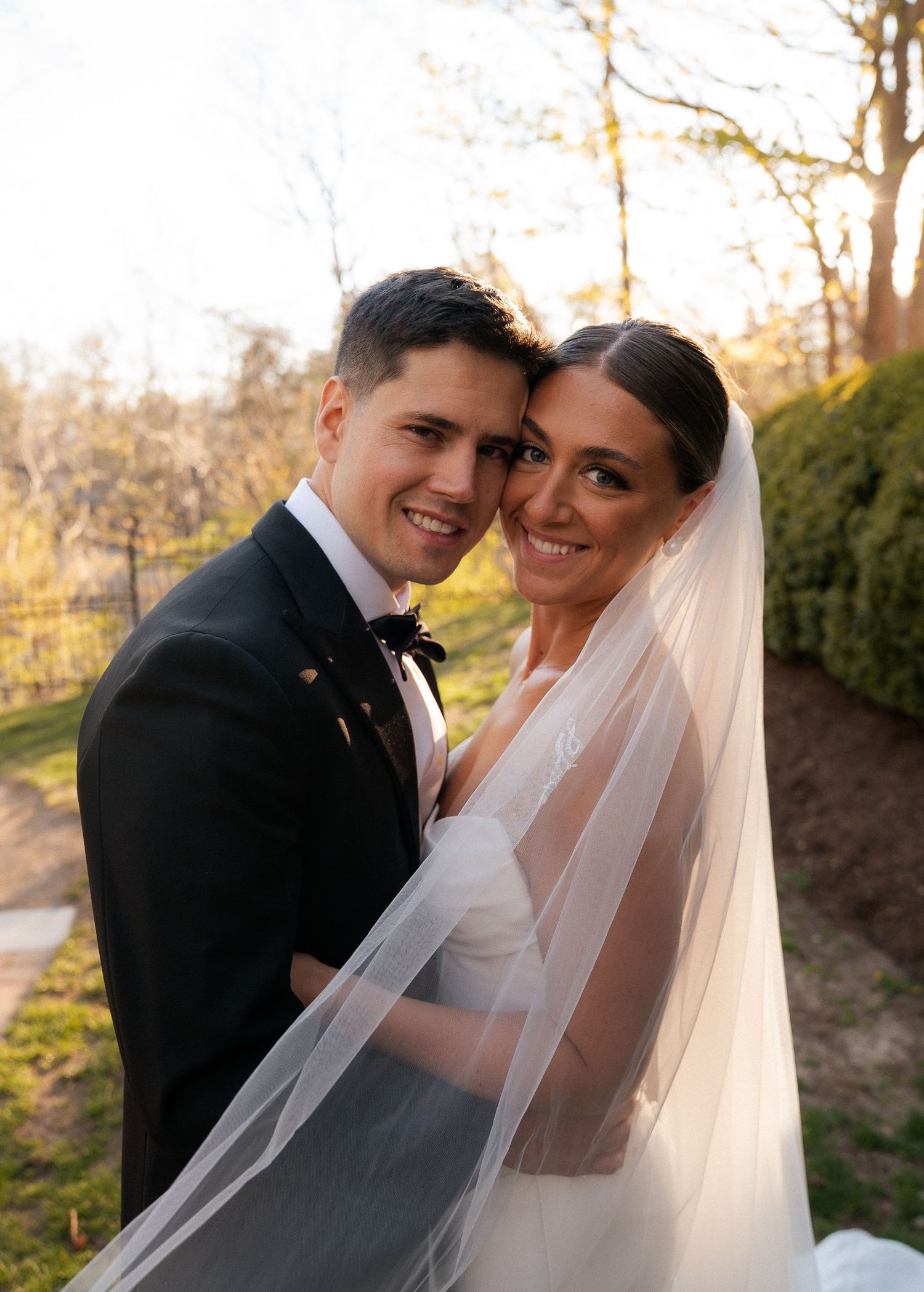 Smiling bride and groom pose closely together in golden hour light with flowing bridal veil.