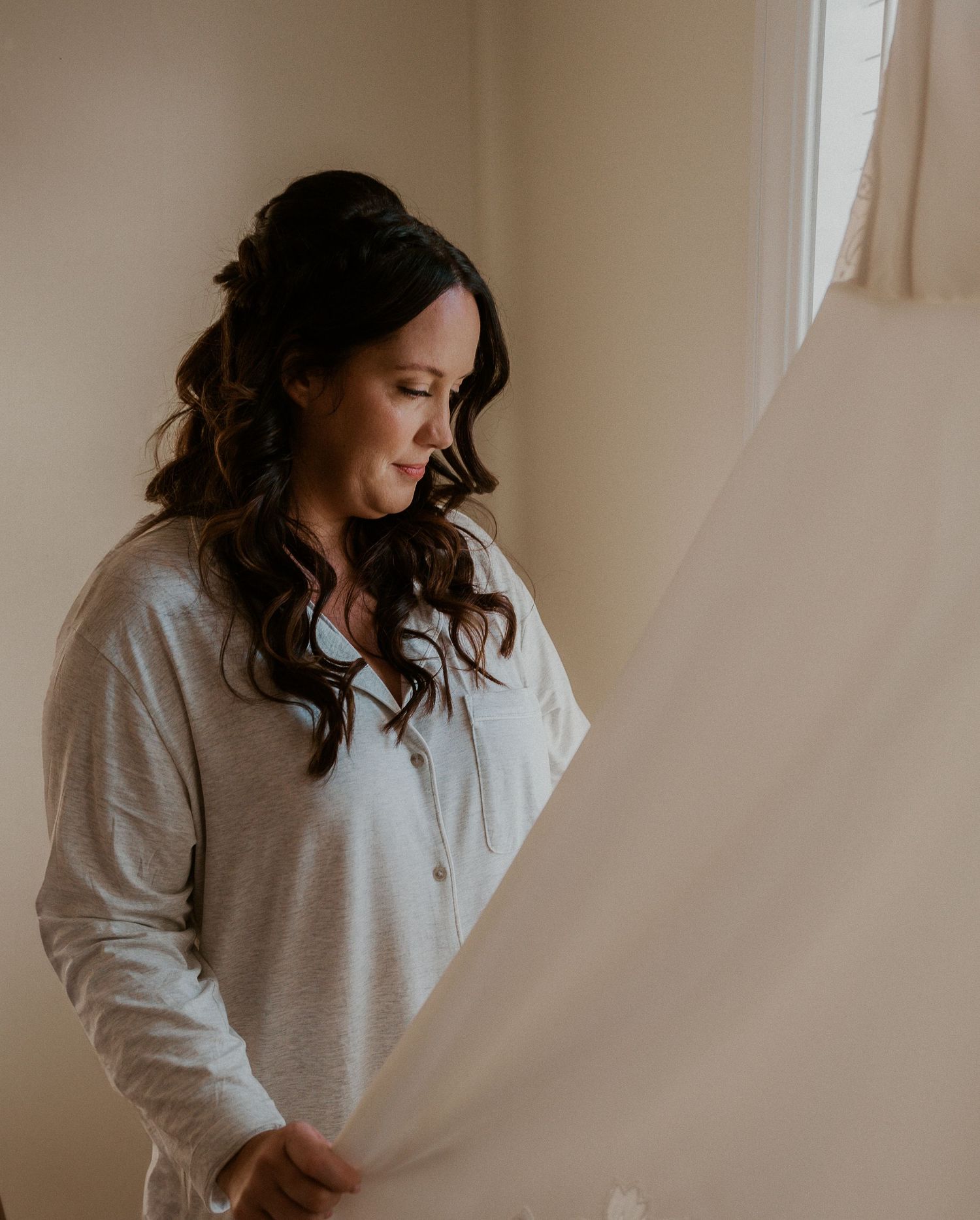 Woman in gray sweater with dark curly hair poses in natural window light for intimate portrait series.