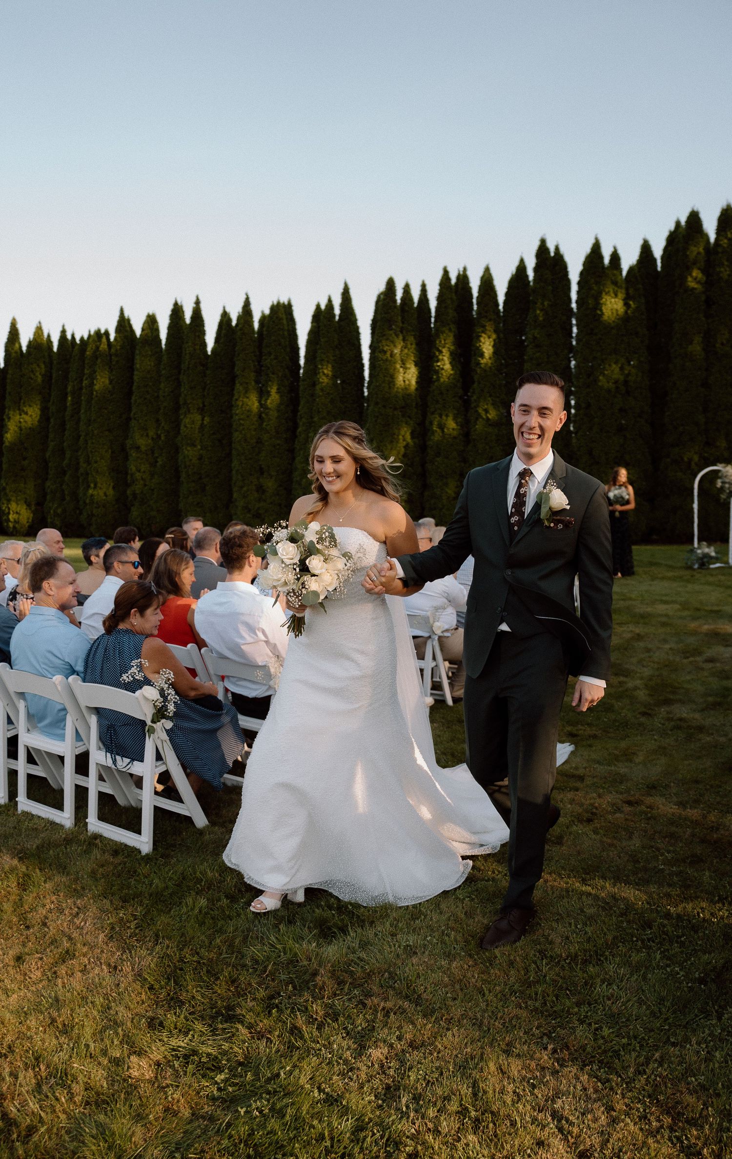 Guests celebrate during a sunset wedding reception on a lawn with evergreen trees.
