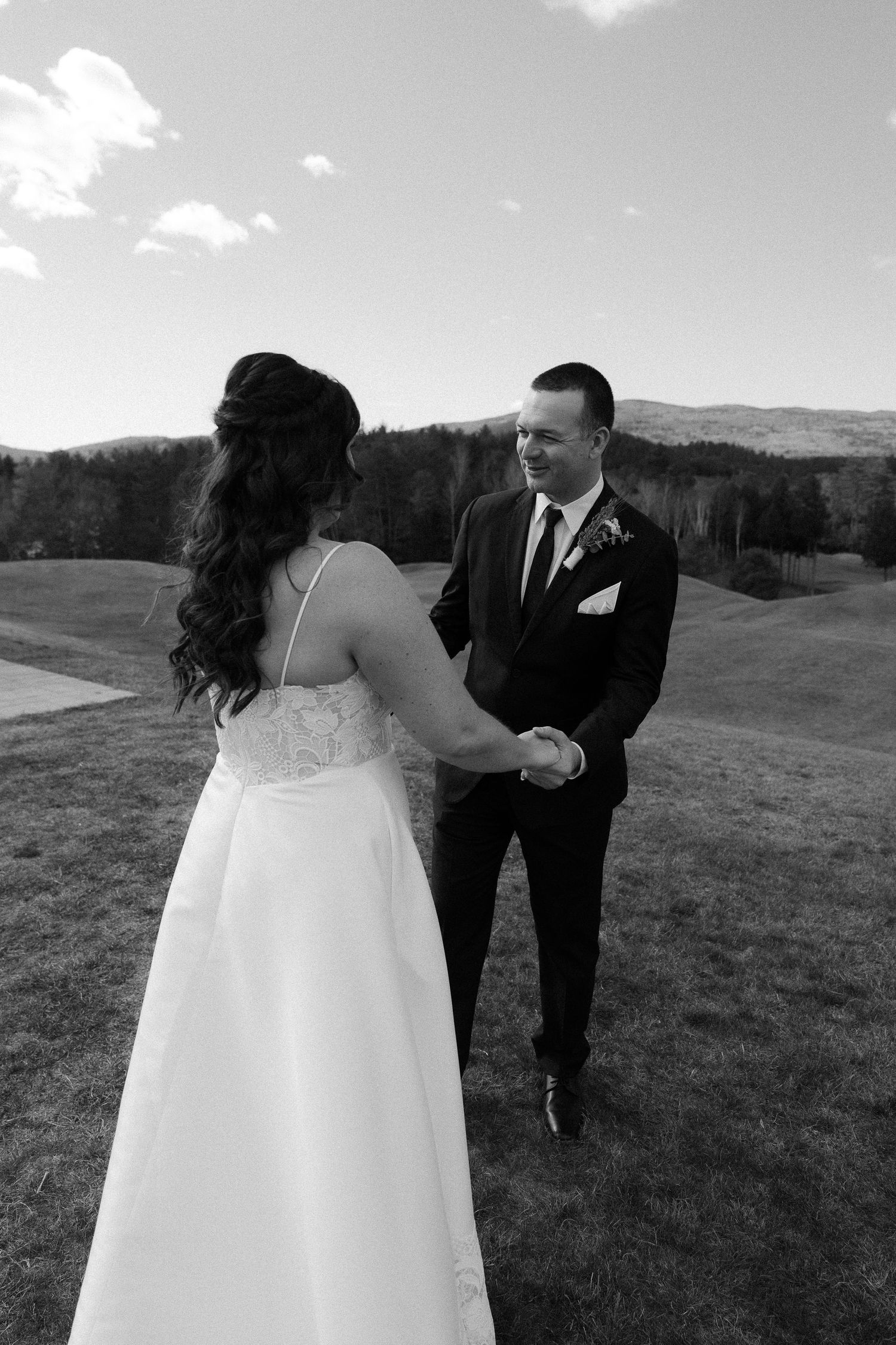 A couple in wedding attire stands together in a grassy field with mountains in the background in black and white.