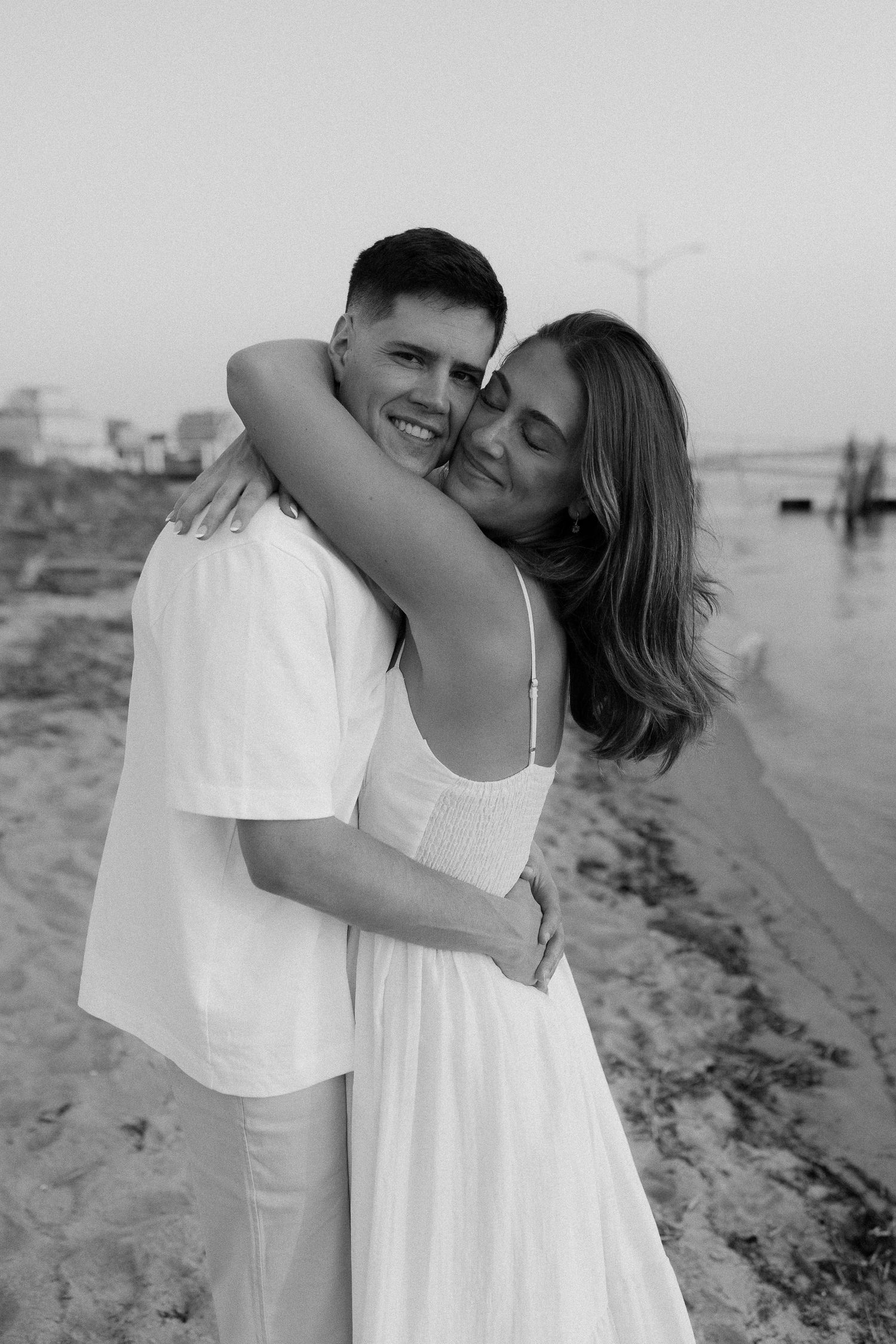Black and white coastal photo of couple embracing in matching white outfits.