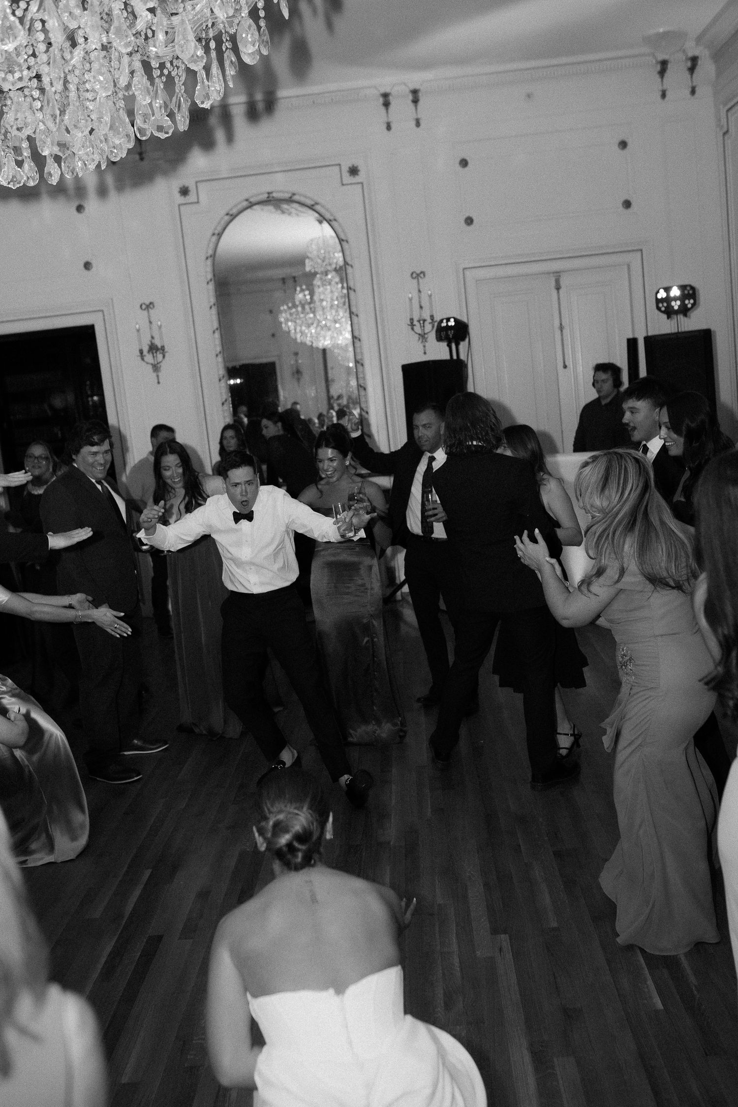 Wedding guests dancing energetically on the dance floor in a grand ballroom with crystal chandeliers.