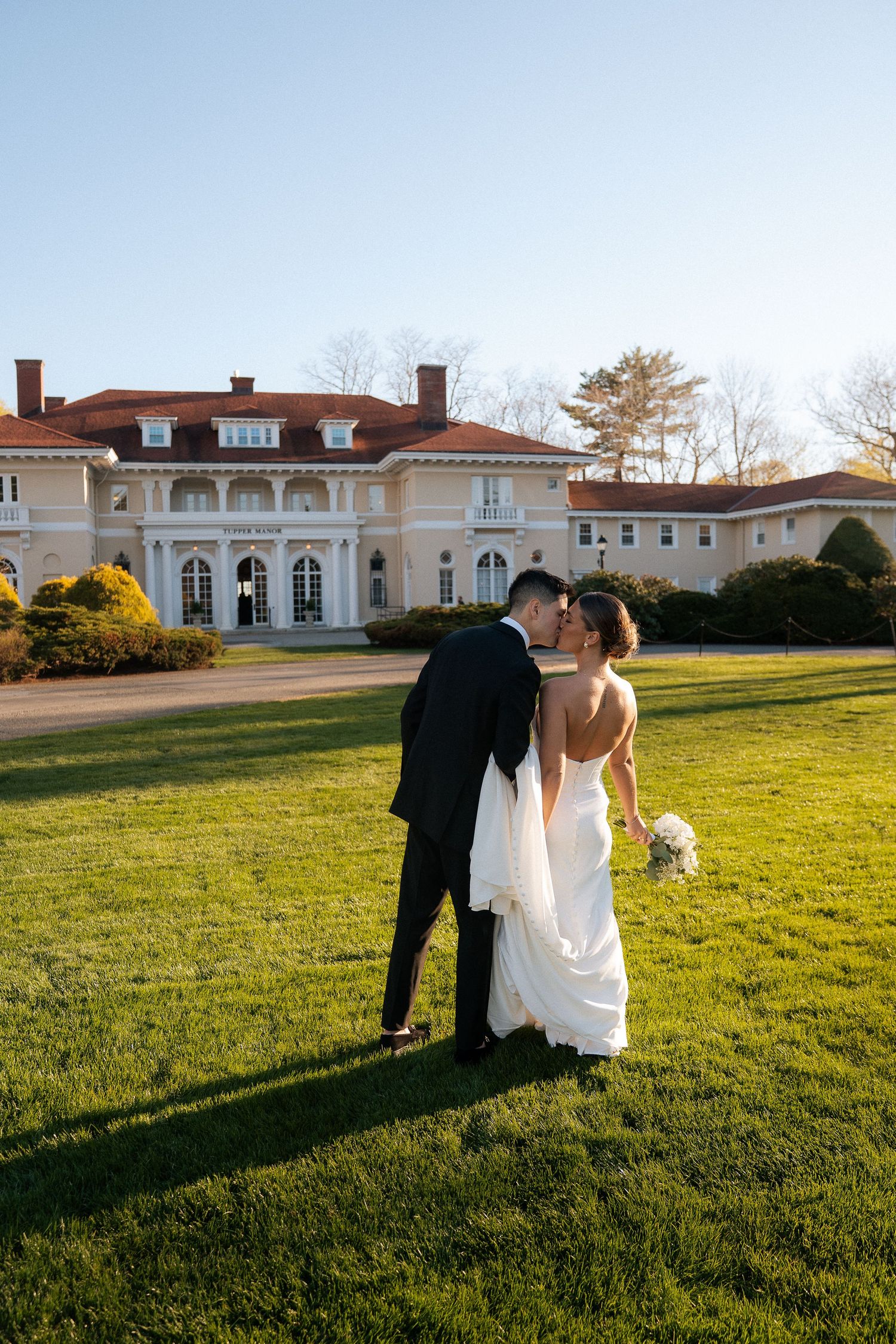 Bride and groom share romantic kiss on sunlit lawn before grand estate wedding venue.