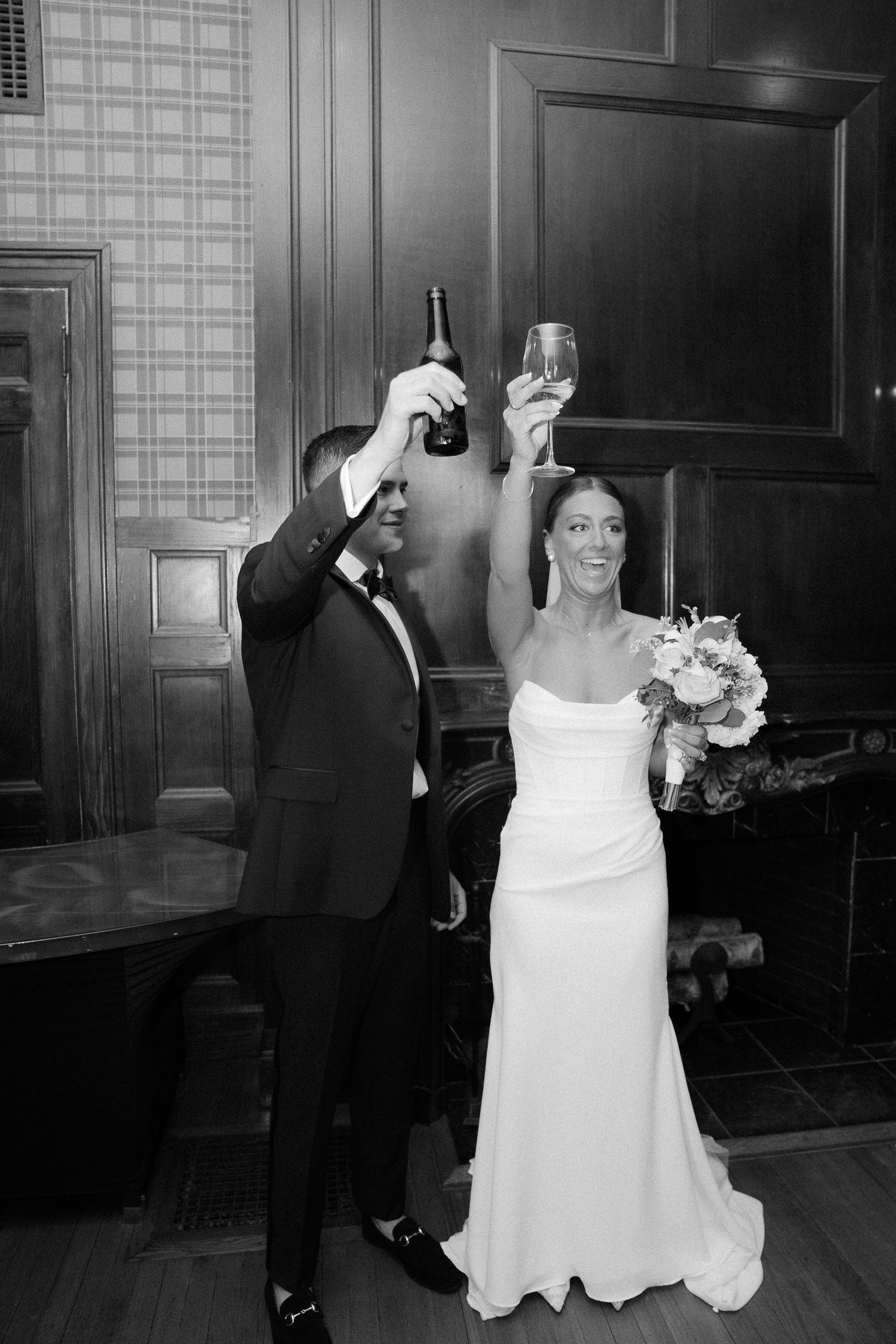 Black and white photo of bride and groom toasting with champagne bottle and glass in a classic wood-paneled room.
