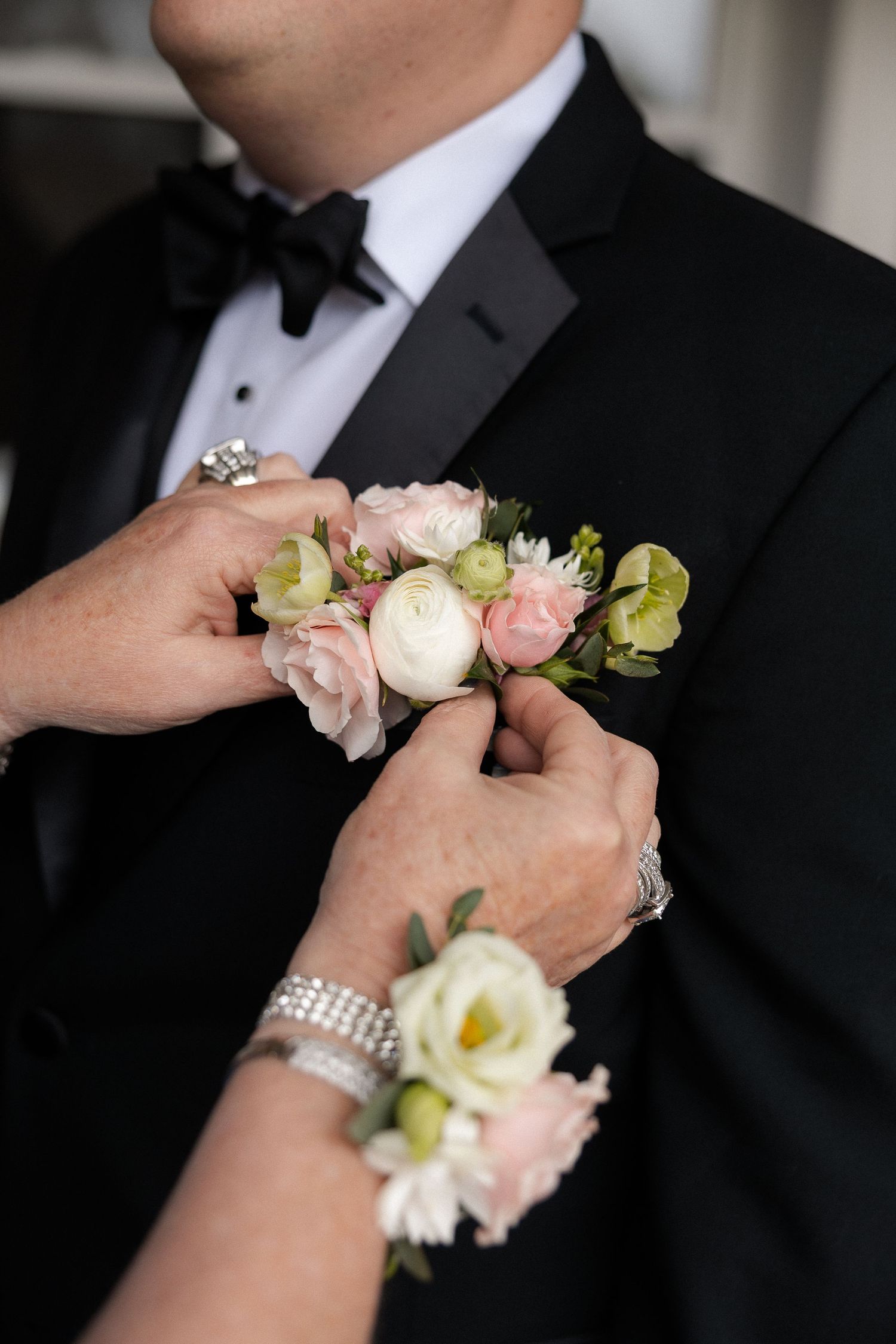 Hands pinning a pink and white floral boutonniere onto a groom's black tuxedo lapel.