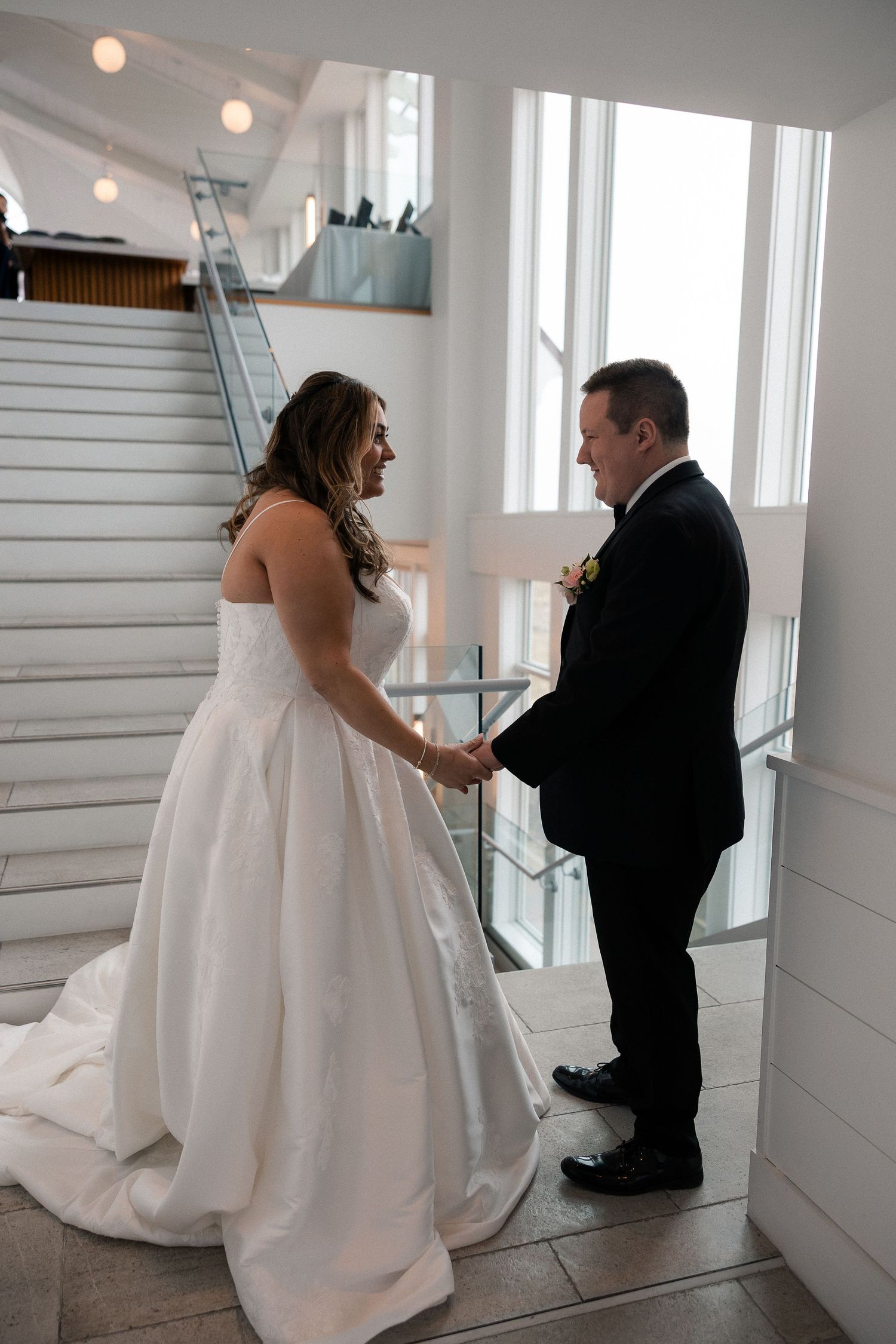 Bride and groom hold hands sharing an emotional first look moment on a staircase in a bright venue.