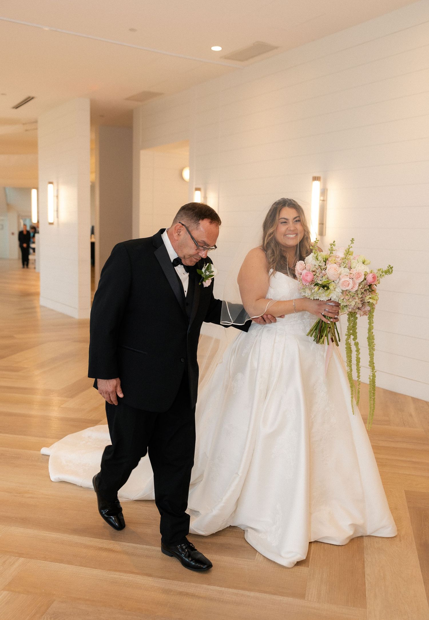 Bride in white ball gown holding pink bouquet walks with man in tuxedo down hallway before wedding ceremony.