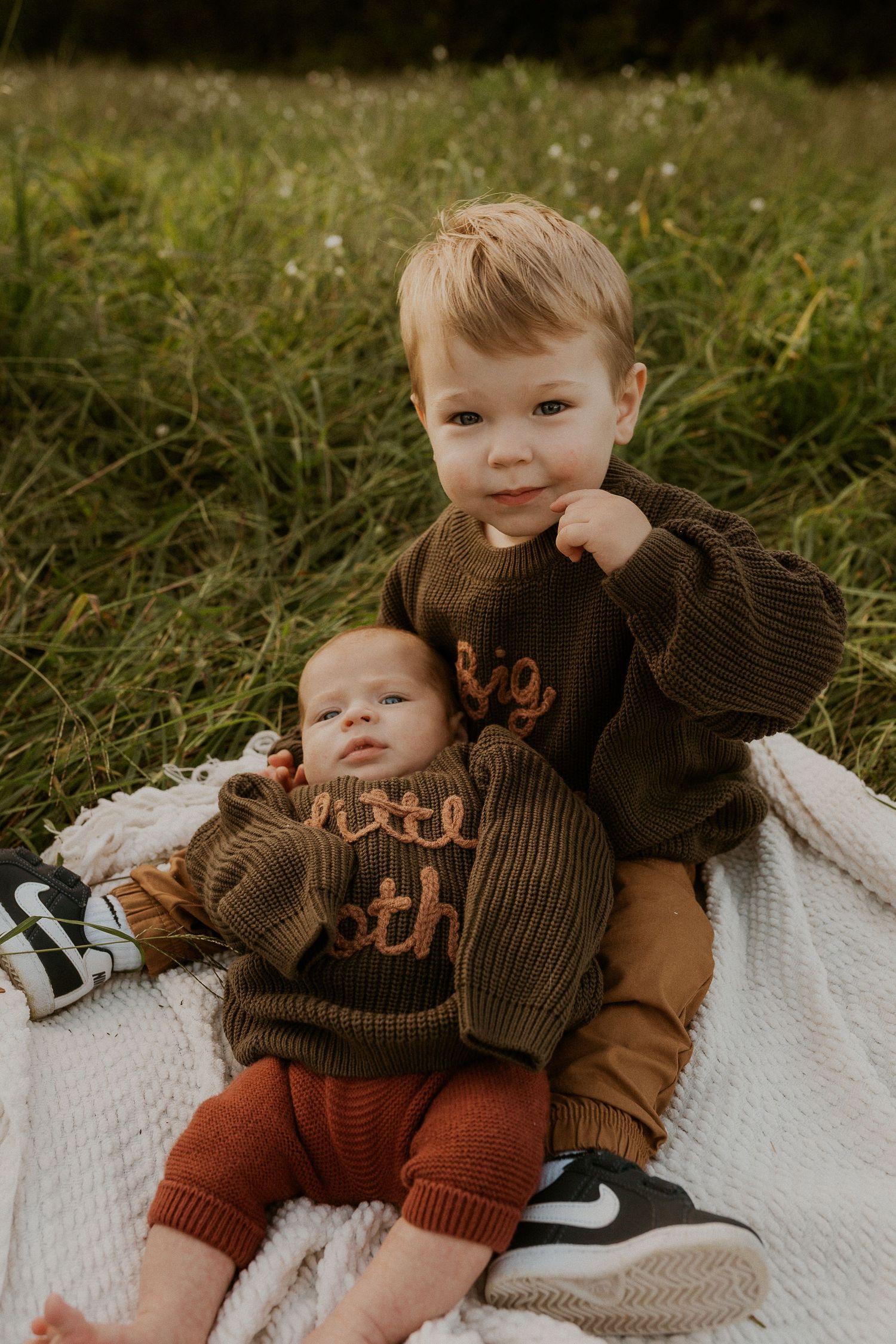 Two siblings in matching brown sweaters share a moment on white blanket.