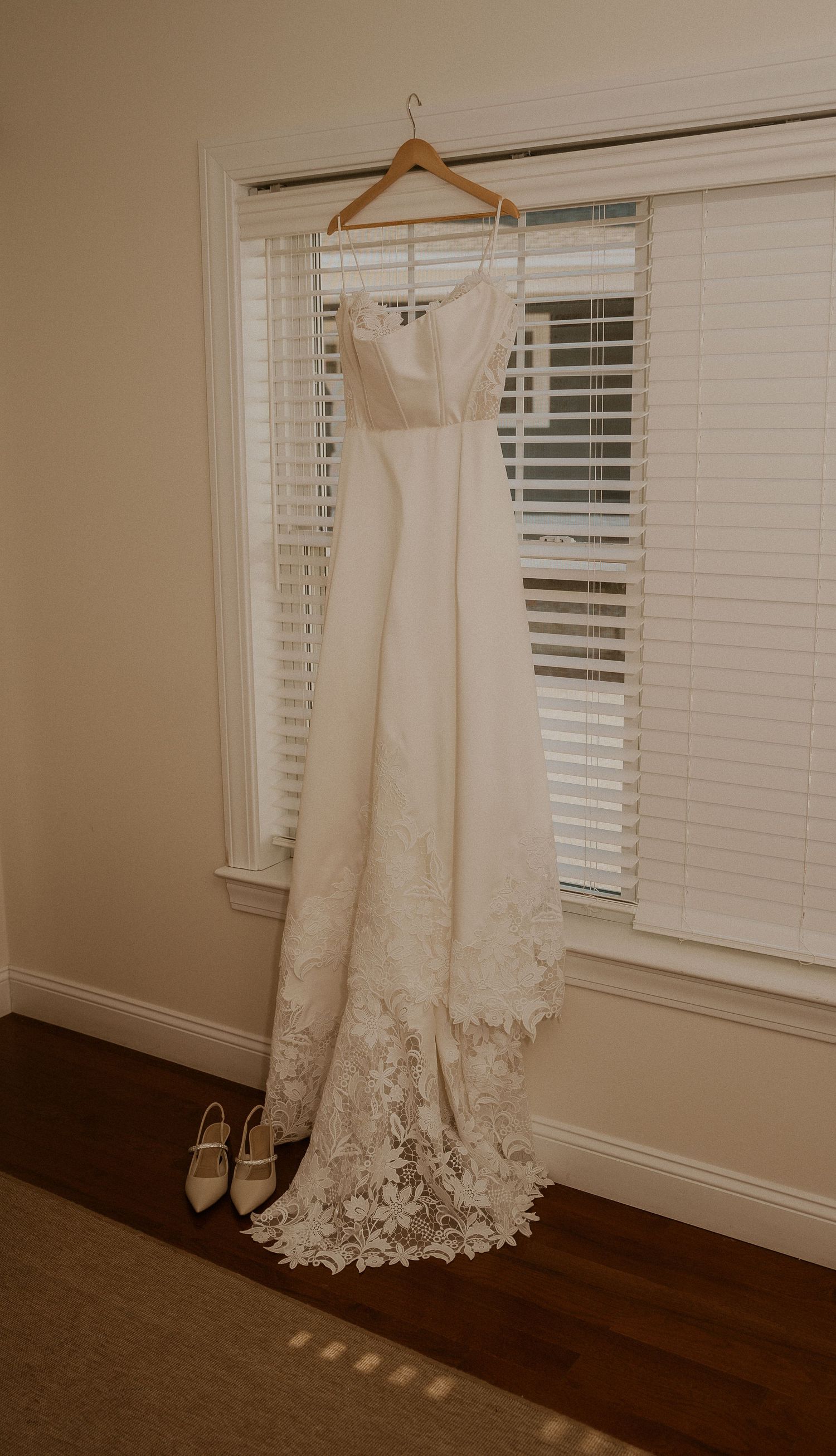 An elegant white wedding dress with lace details hangs by a window with shoes beneath.