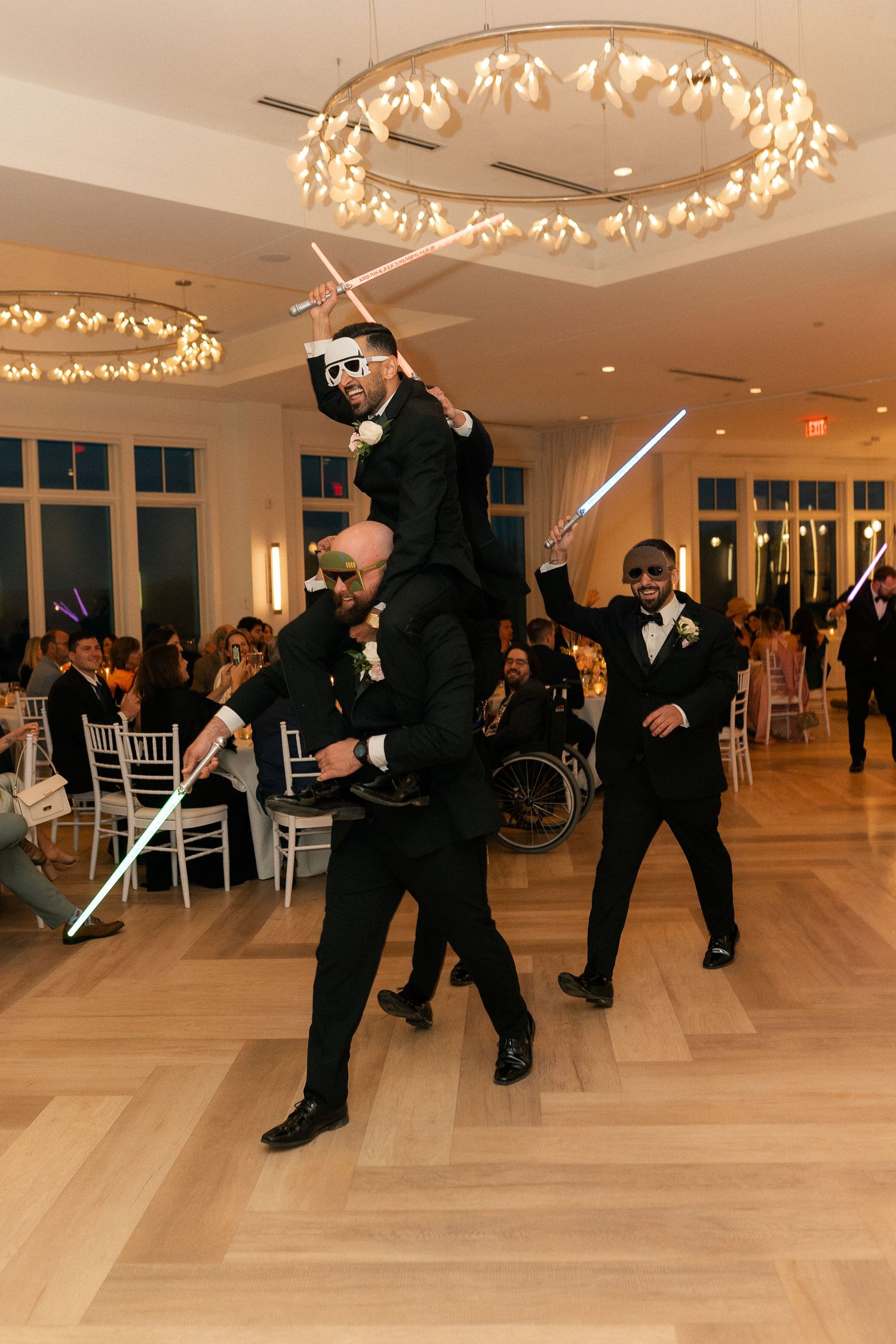 Groomsmen in black tuxedos celebrate with light sabers on dance floor at wedding reception.