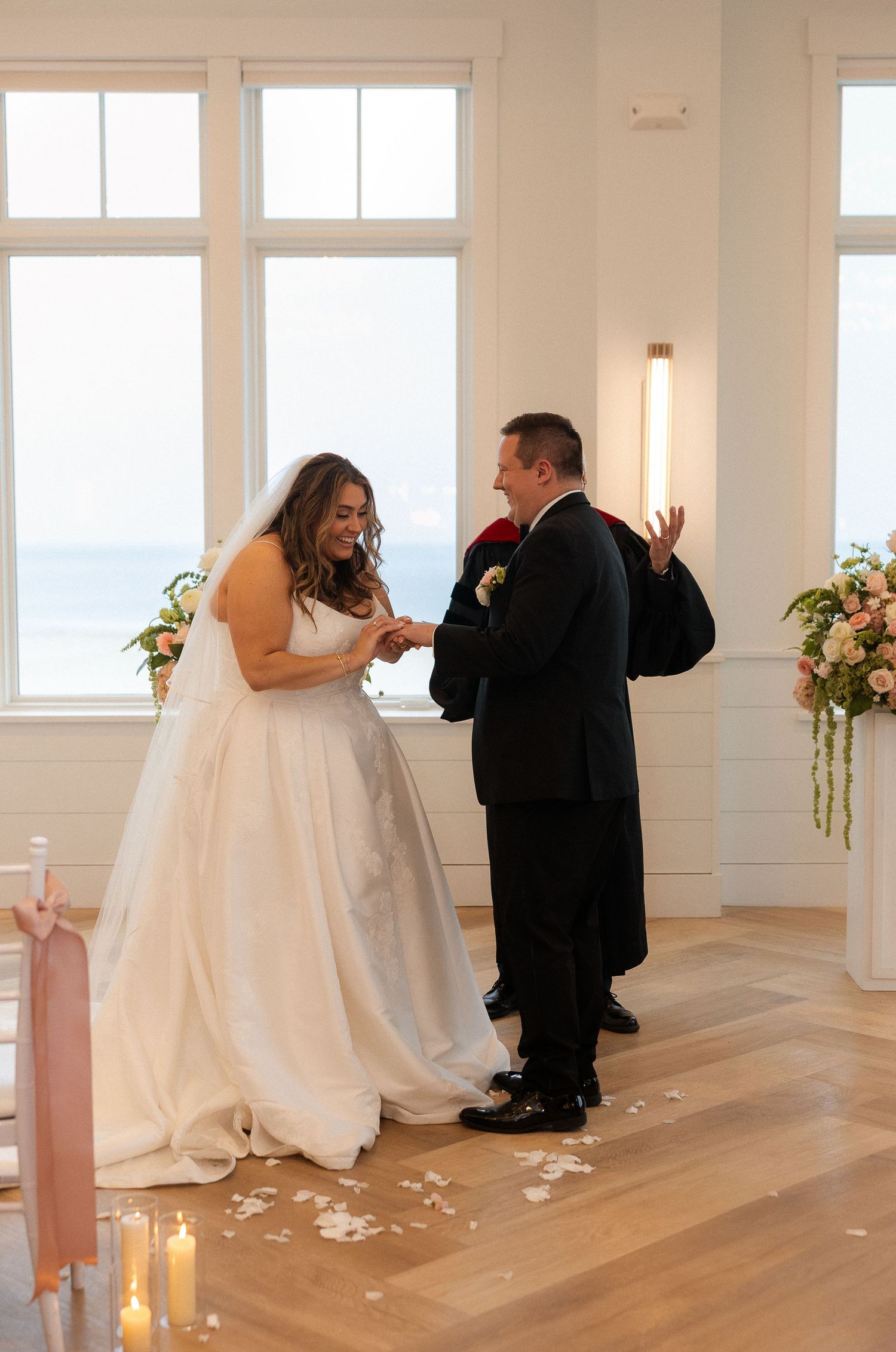 Bride in white gown exchanges rings with groom at oceanview altar with floral arrangements and candles.