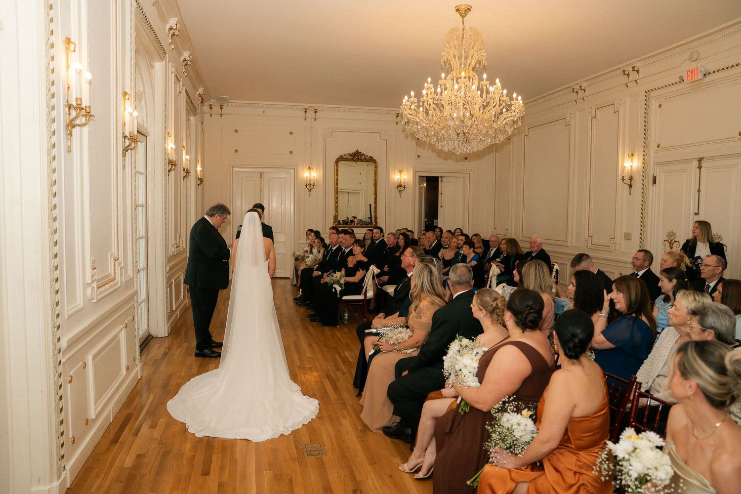 Elegant wedding ceremony in ornate ballroom with chandelier, bride in white gown at altar with seated guests in formal attire.