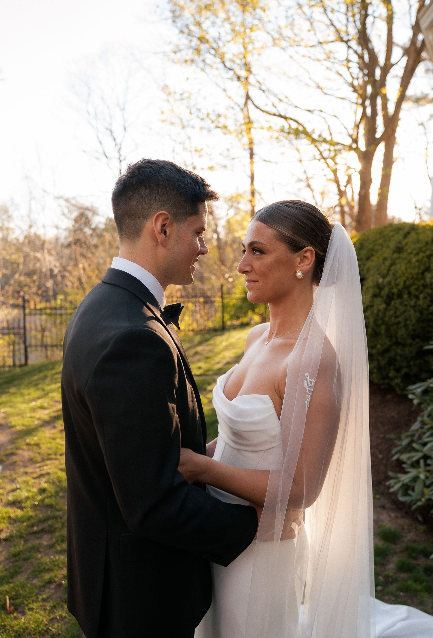 Bride and groom gazing at each other in garden at golden hour, bride wearing strapless gown and veil.
