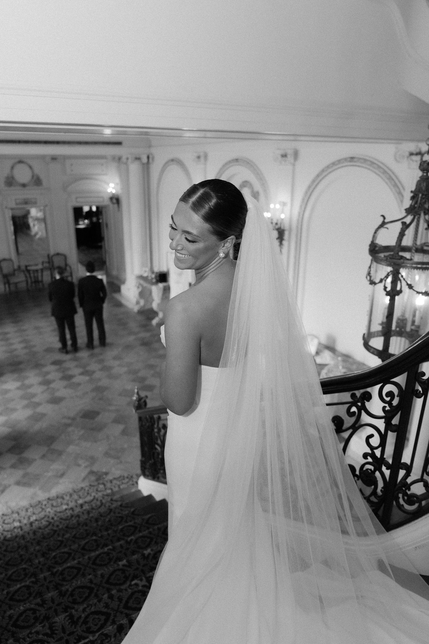 Black and white photo of a smiling bride in a strapless gown and long veil on an elegant staircase.
