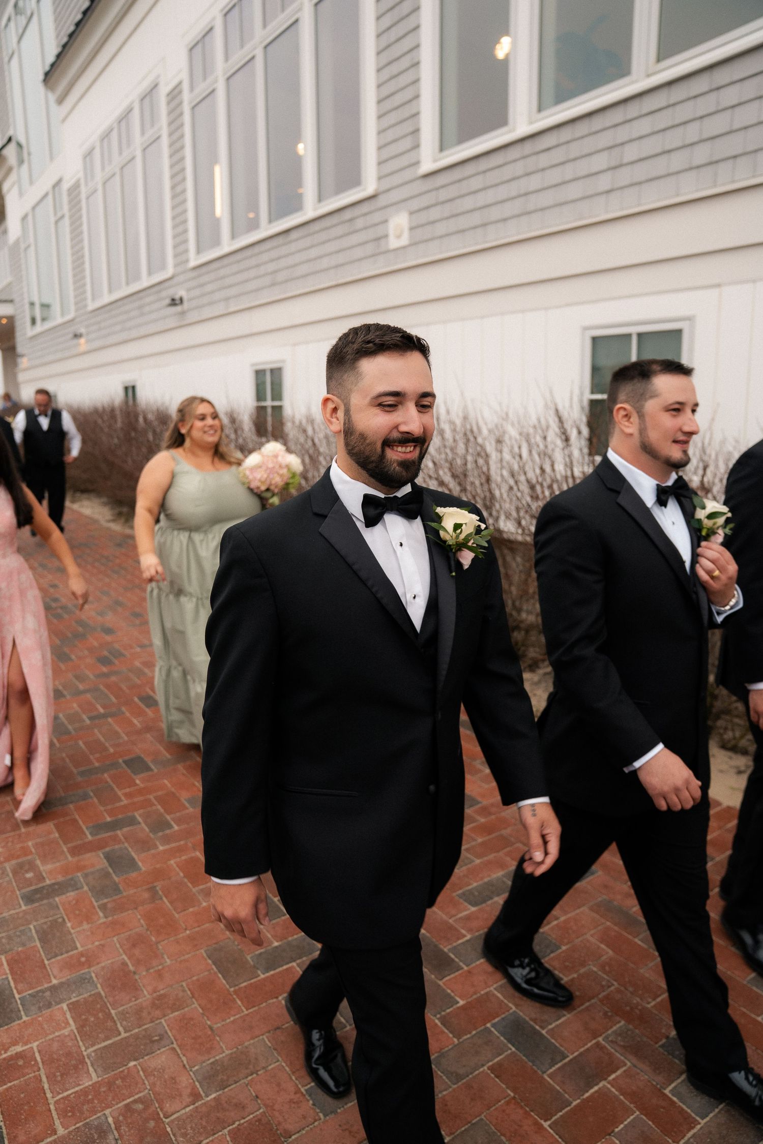 Smiling groom in black tuxedo walks with groomsmen outside wedding venue on brick pathway.