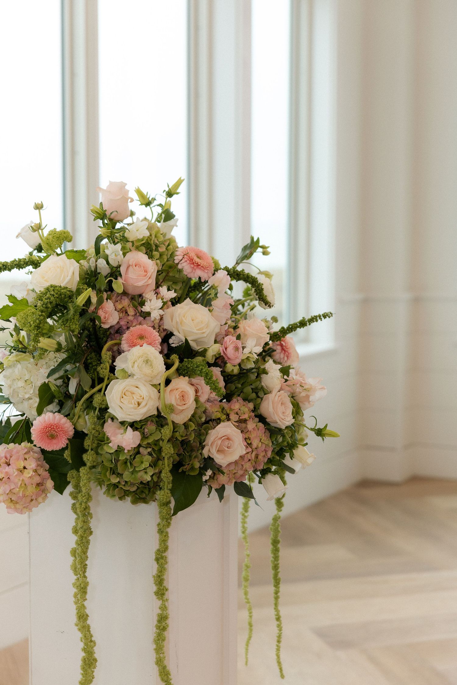 Lush wedding floral arrangement with pink roses, white blooms, and cascading greenery on a white pedestal.