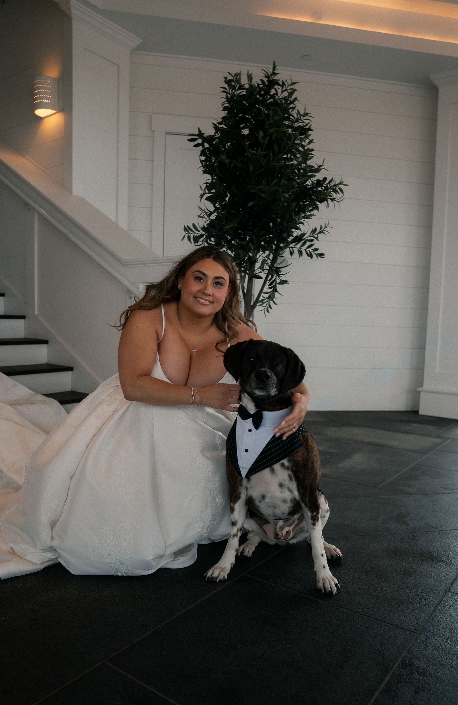 Bride in white gown posing with dog dressed in tuxedo bow tie on dark tile floor near white staircase.