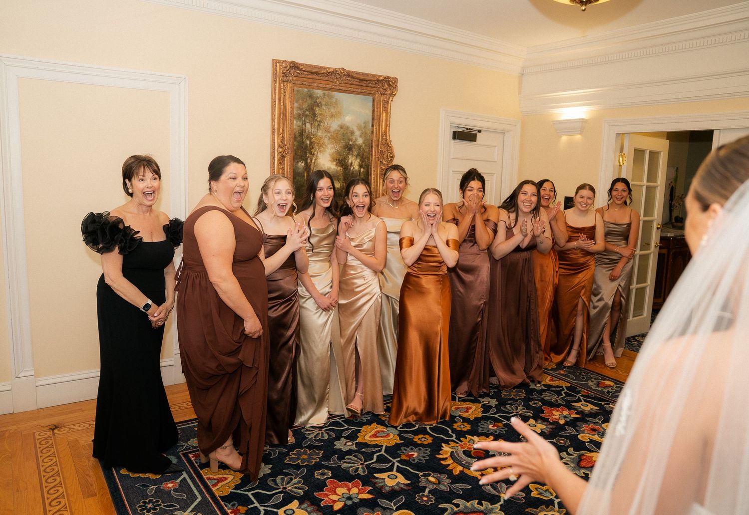 Bridesmaids in brown and gold dresses react with joy as bride in white veil reveals her wedding look indoors.