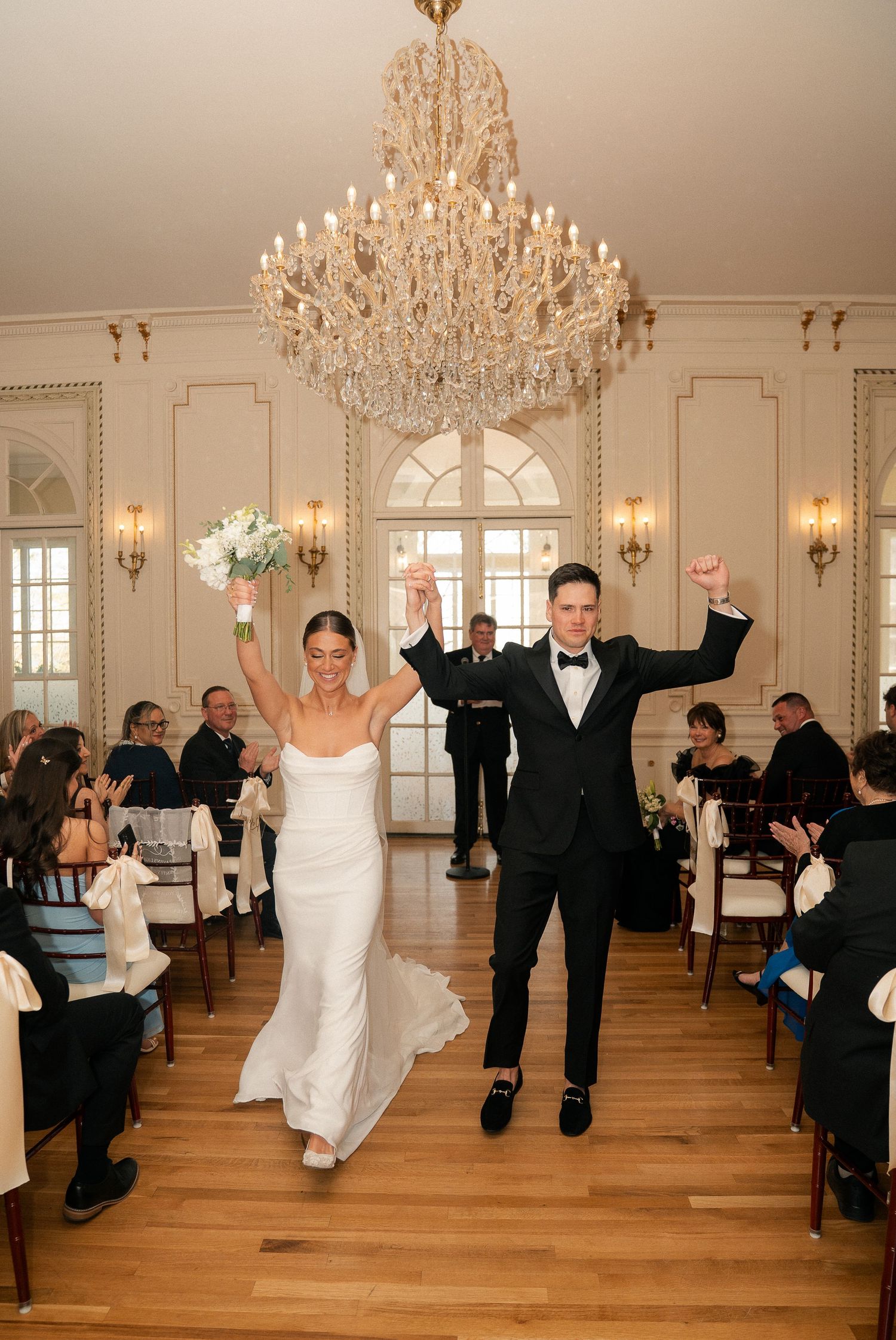 Bride and groom celebrate their wedding ceremony exit with raised arms under a crystal chandelier in an elegant ballroom.